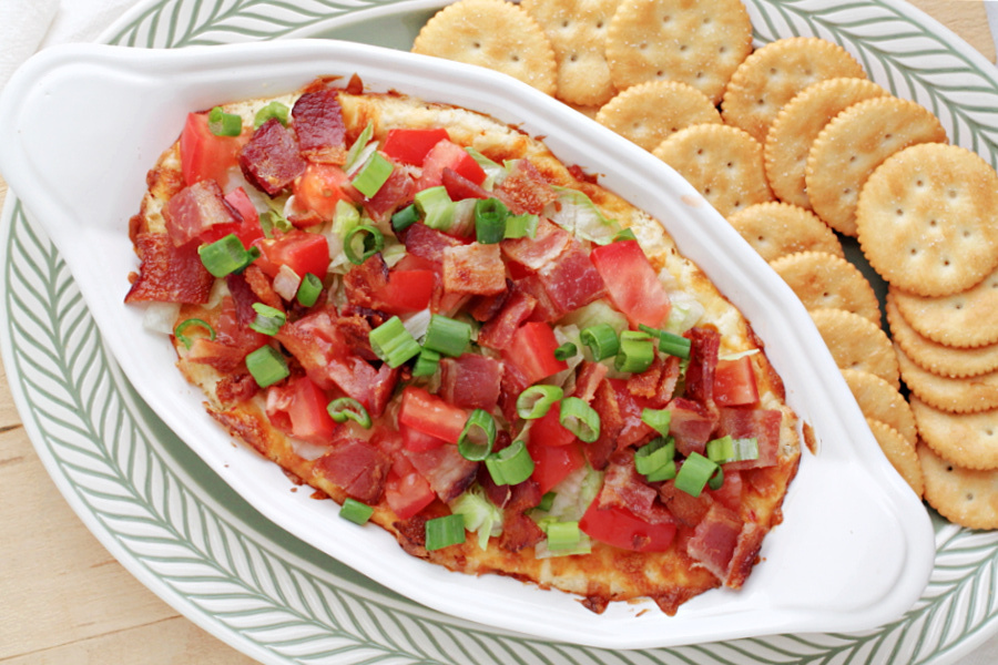 BLT dip in an oval dish, topped with diced tomatoes, crispy bacon, and fresh green onions, surrounded by round crackers on a patterned plate.