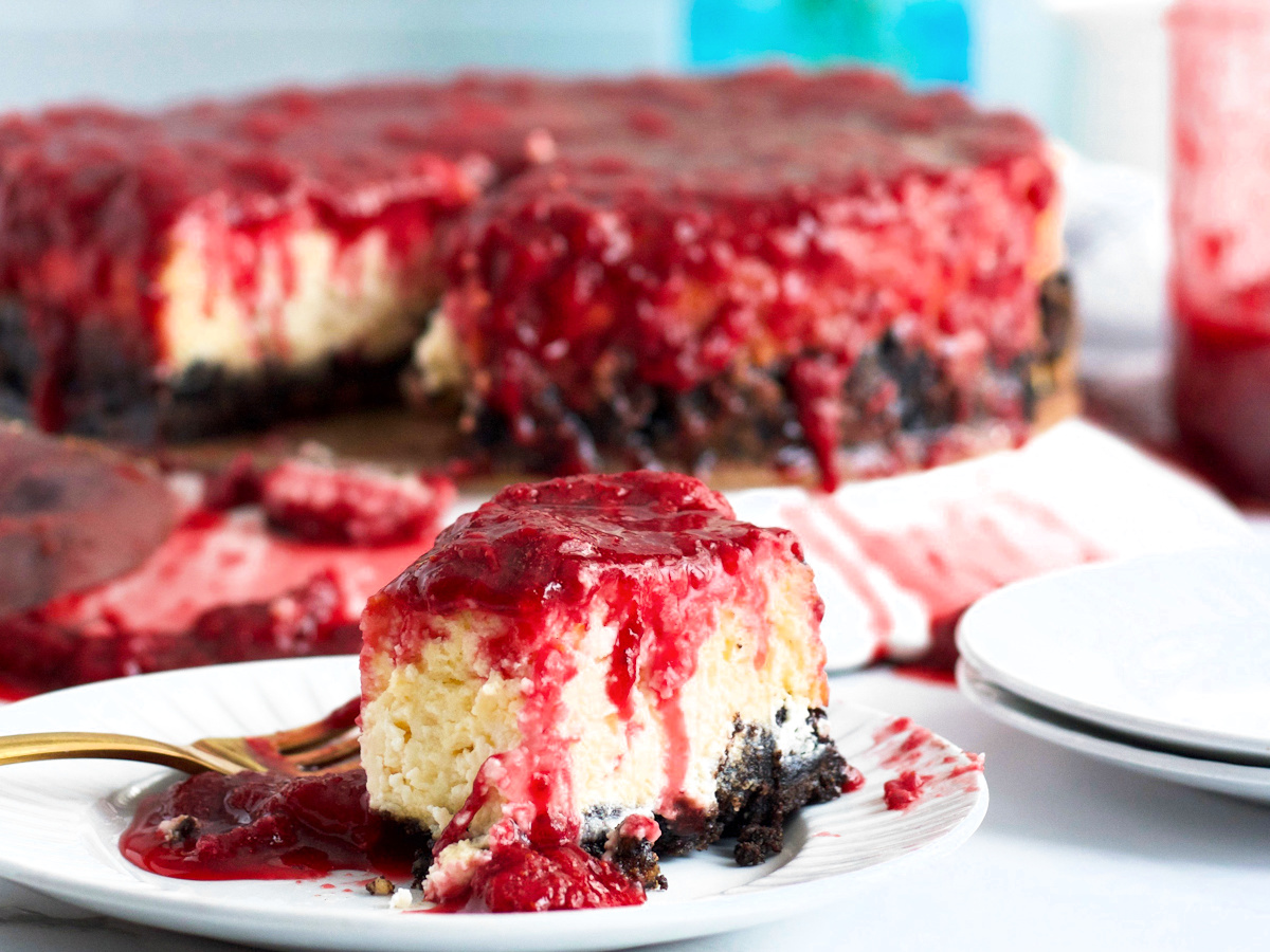 A slice of classic strawberry cheesecake with Oreo crust gleams with raspberry topping on a plate, surrounded by the larger cheesecake from which it was cut. A fork lies invitingly next to the delectable slice.