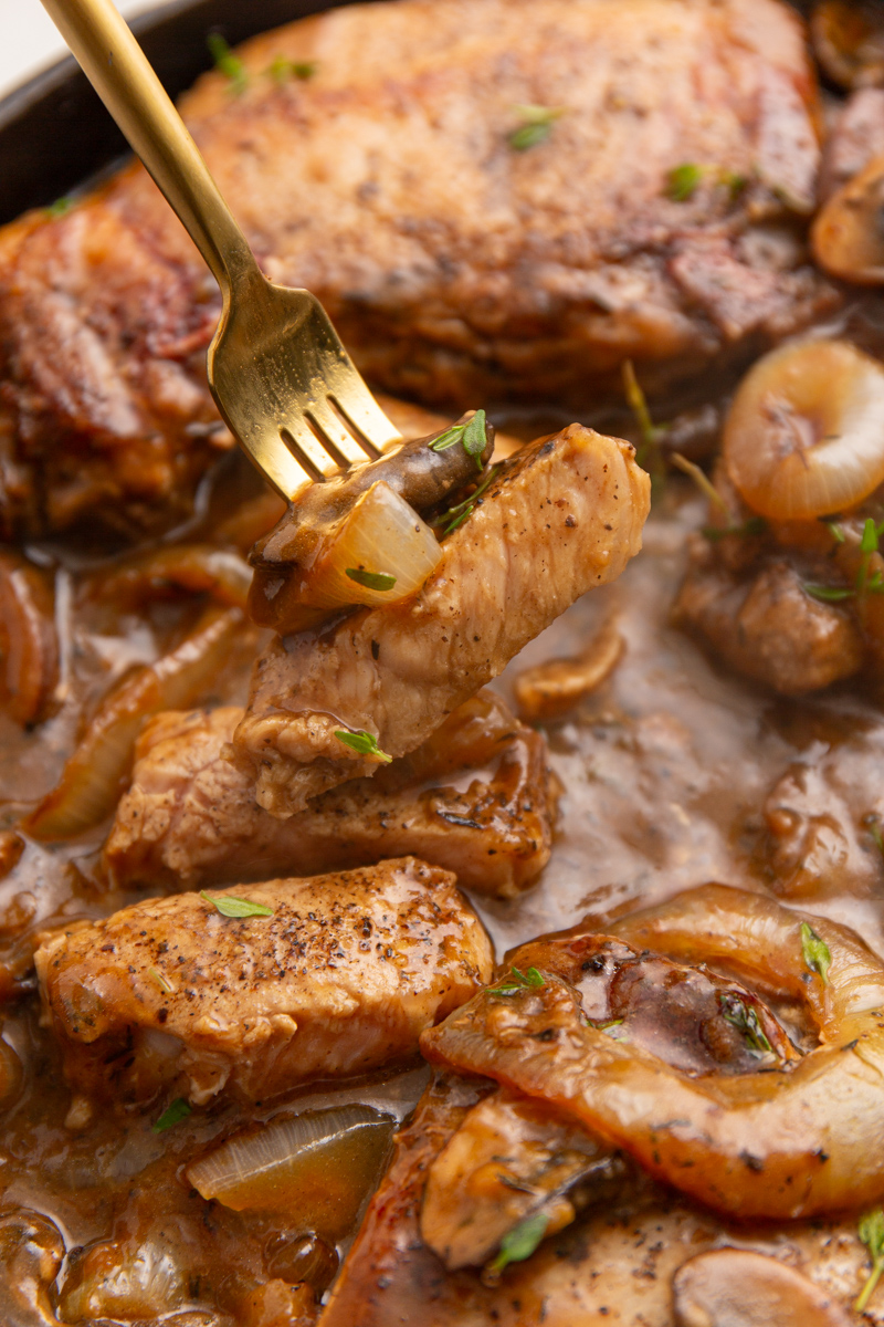 Close-up of a fork lifting a piece of Salisbury pork chop from a skillet with cooked onions and mushrooms in a rich brown sauce.