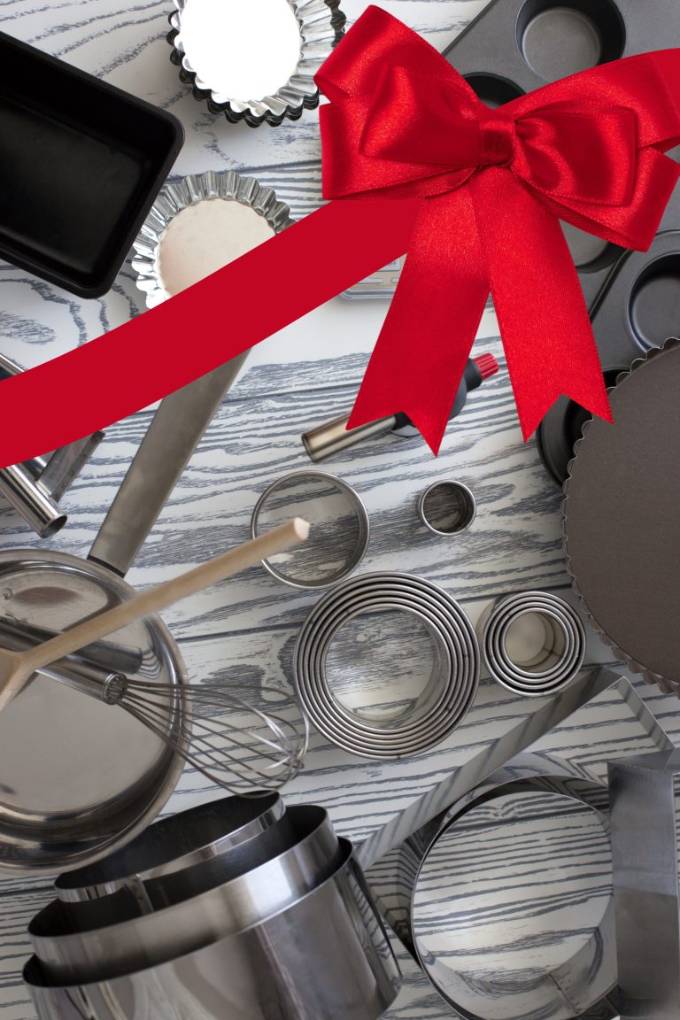 A collection of baking tools, perfect gifts for bakers, includes pans, cookie cutters, and a whisk. They're artfully arranged on a wood-grain surface with a large red ribbon across the top.