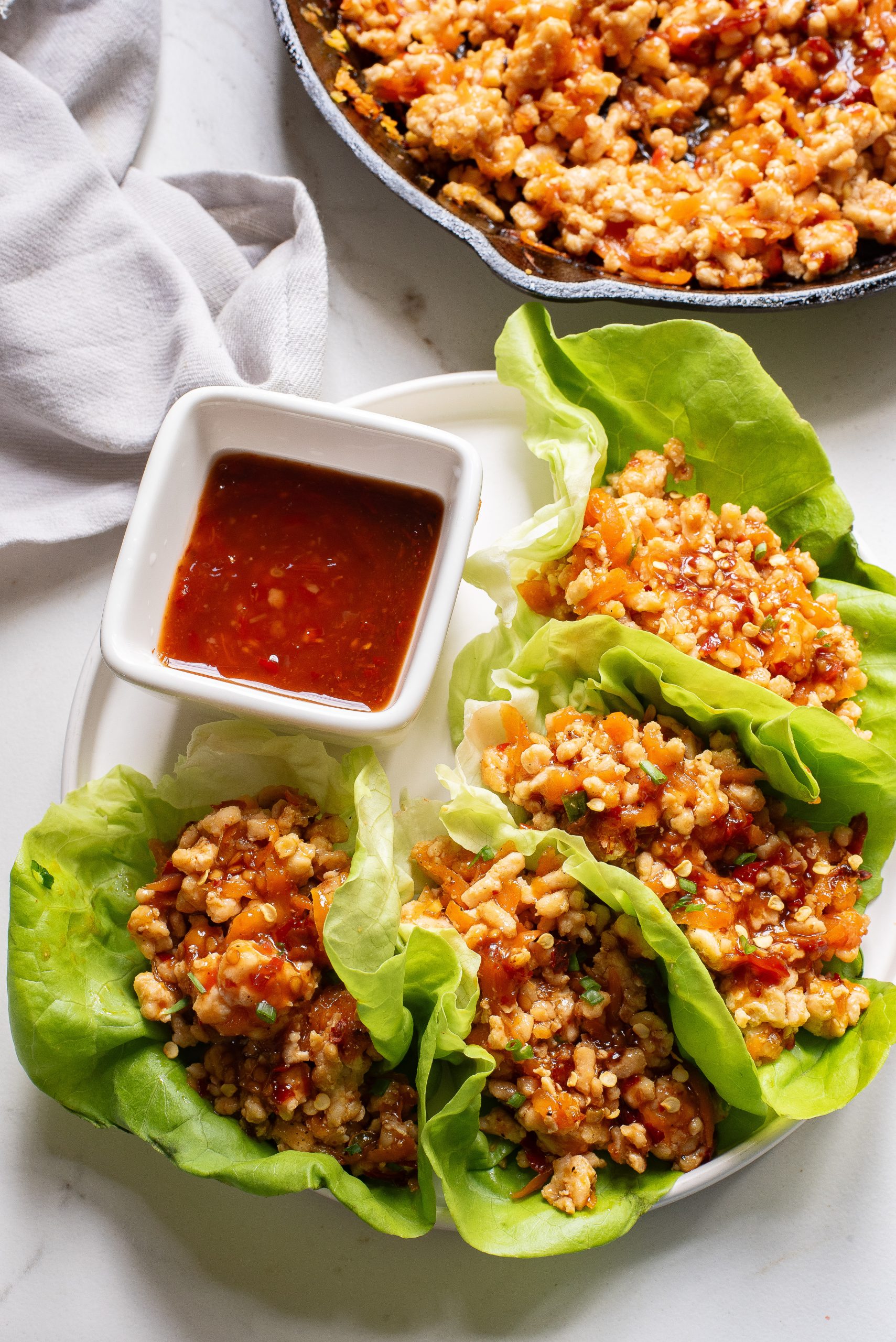 A plate of chicken lettuce wraps with a side of spicy sauce, served alongside a skillet of extra filling.