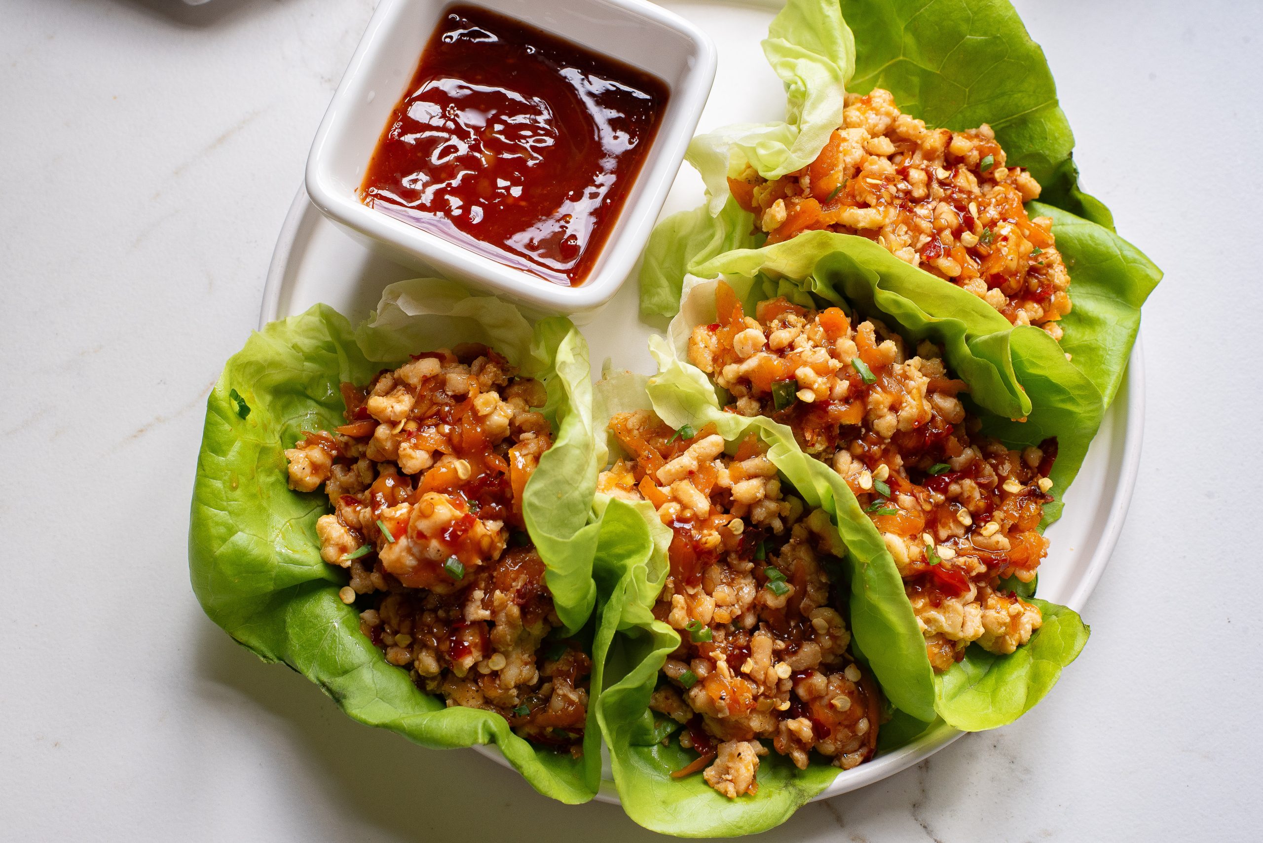 Lettuce wraps filled with savory minced meat and grains, served with a side of red sauce on a white plate.