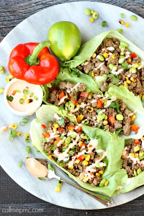 Two Keto Asian Chicken Lettuce Wraps filled with ground beef, corn, edamame, and diced red bell peppers, served on a plate with a whole lime, a red bell pepper, chopped green onions, and a side of dipping sauce.