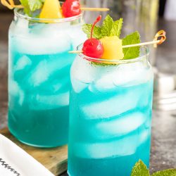 Two glasses of Blue Hawaiian Cocktail with ice, garnished with cherries, pineapple chunks, and mint leaves, are placed on a table with a white napkin nearby.