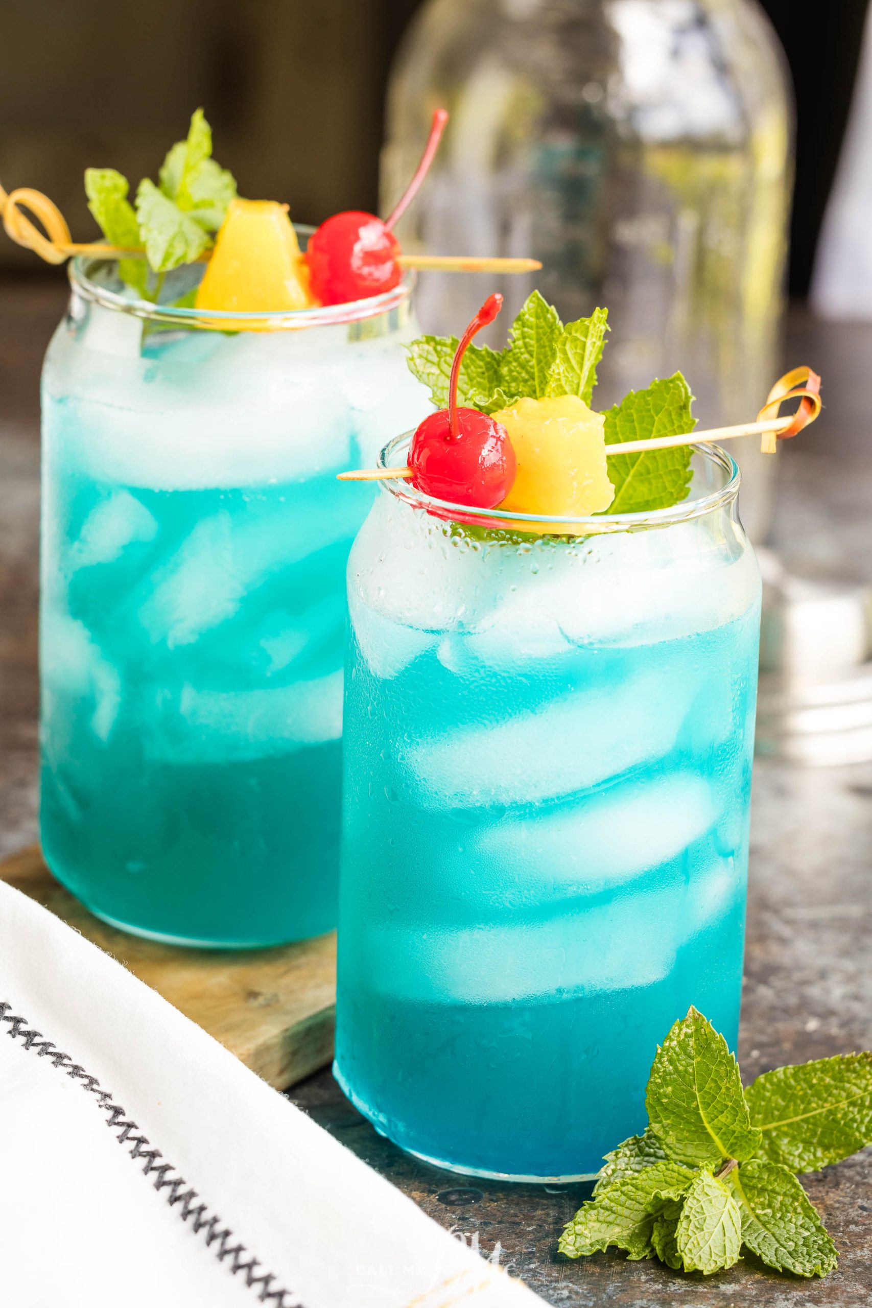Two glasses of Blue Hawaiian Cocktail with ice, garnished with cherries, pineapple chunks, and mint leaves, are placed on a table with a white napkin nearby.