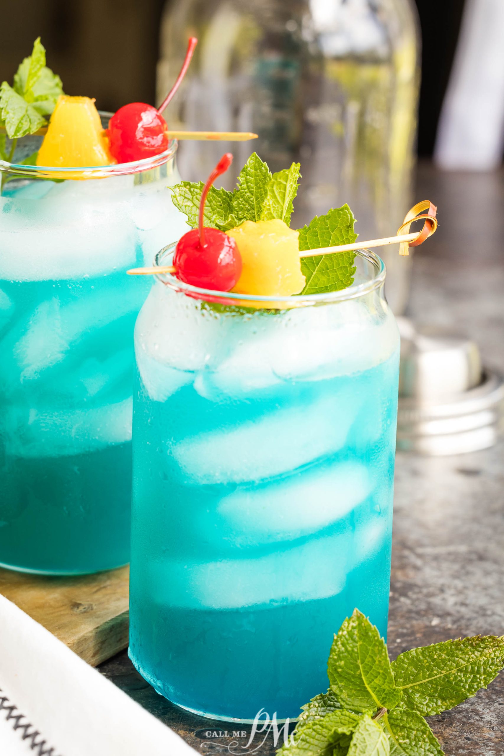Two glasses of blue cocktails with ice, garnished with mint leaves, a maraschino cherry, and a pineapple chunk, are placed on a wooden board.