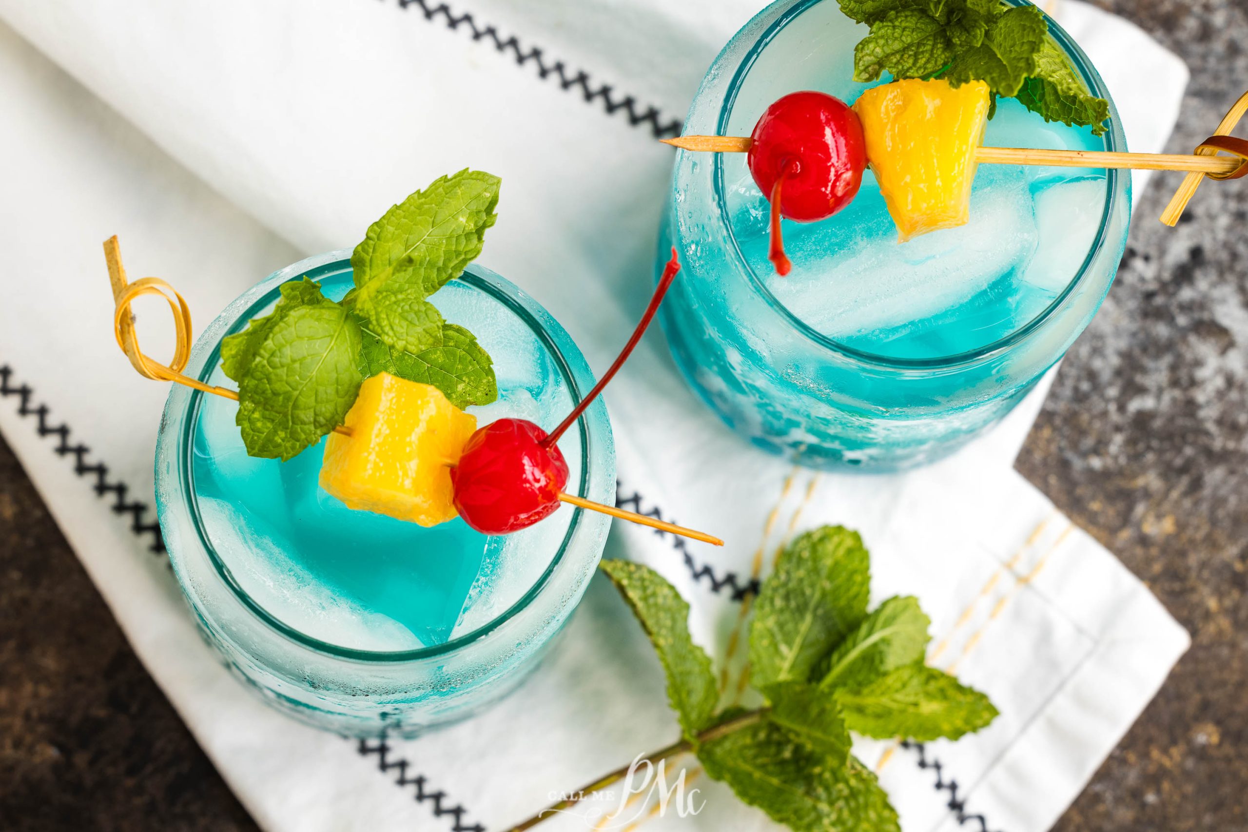 Two drinks with ice, garnished with skewers of a red cherry, yellow pineapple chunk, and fresh mint, placed on a white cloth with black stitching. Mint leaves are scattered nearby.