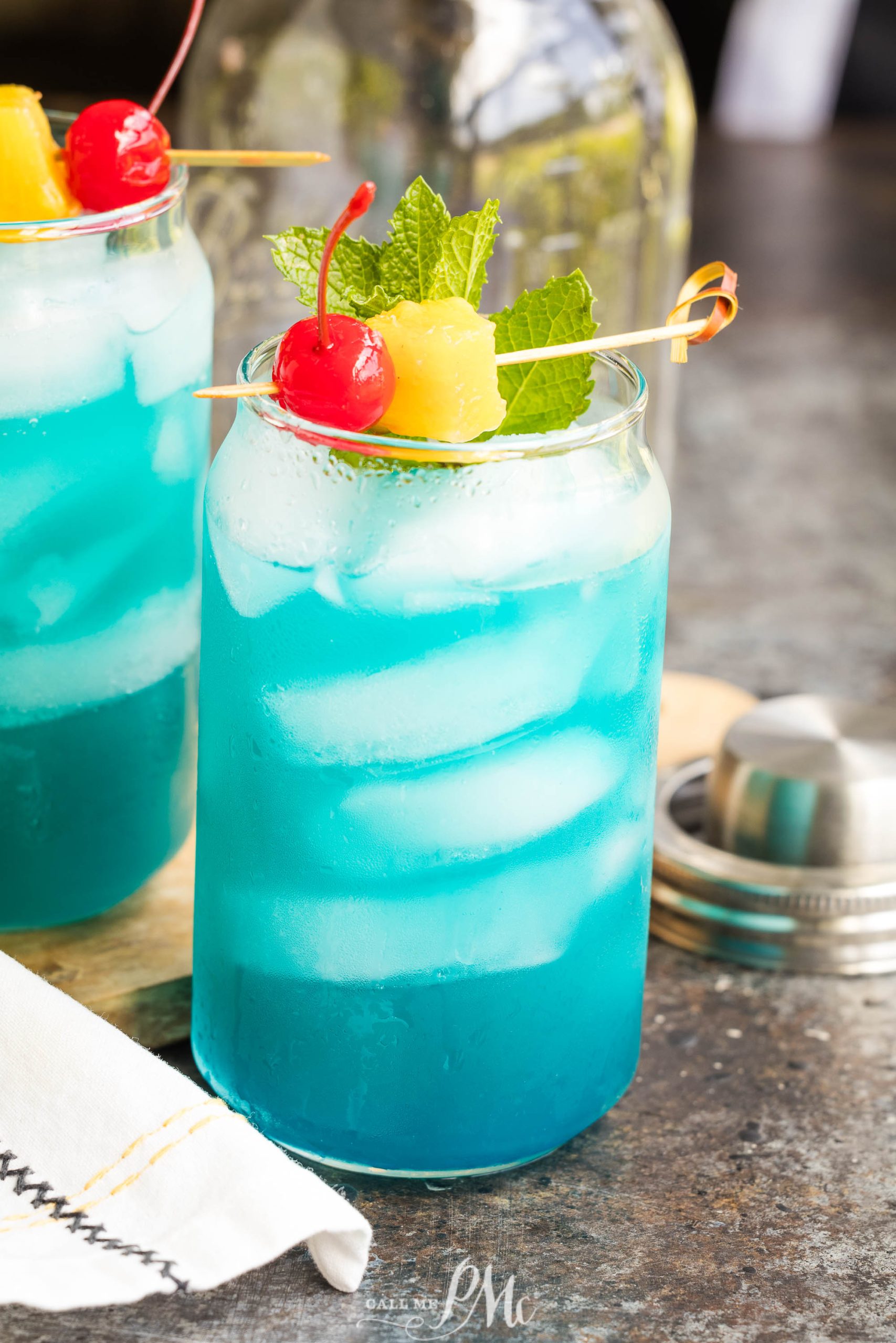 A clear glass filled with a blue liquid, ice cubes, and garnished with a cherry, pineapple chunks, and mint leaves on a skewer.