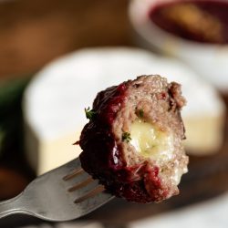 A fork holding a bite of meat with oozing cheese and a berry sauce, with a whole round cheese and bowl of sauce blurred in the background.