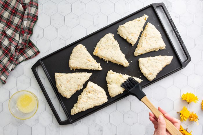A hand brushes the tops of unbaked triangular Cheddar Herb Scones on a black baking tray with a melted butter mixture. Nearby are a cloth towel, a small bowl with butter, and yellow flowers on a hexagonal tile surface.