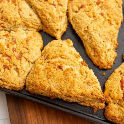 A tray of six golden-brown scones, arranged in neat, spaced intervals on a black baking sheet, rests on a wooden cutting board.