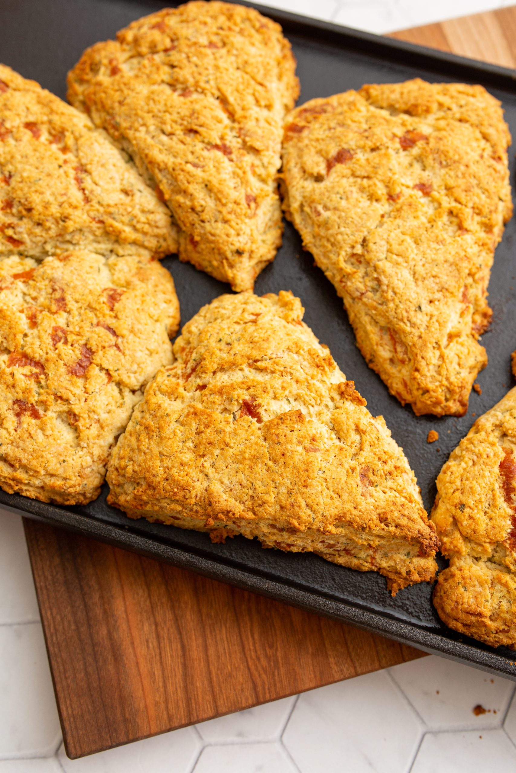 A tray of six golden-brown Cheddar Herb Scones, arranged in neat, spaced intervals on a black baking sheet, rests on a wooden cutting board.