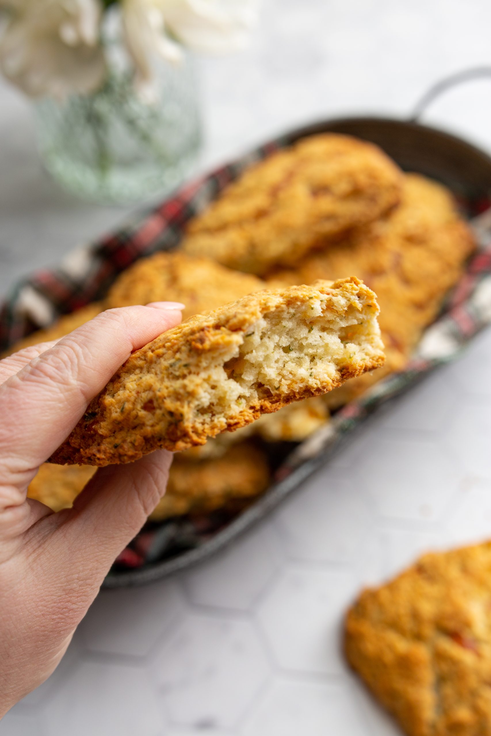 Close-up of a hand holding a half-eaten Cheddar Herb Scones, showing the crumbly interior. A tray filled with more scones and a vase of white flowers are in the background.