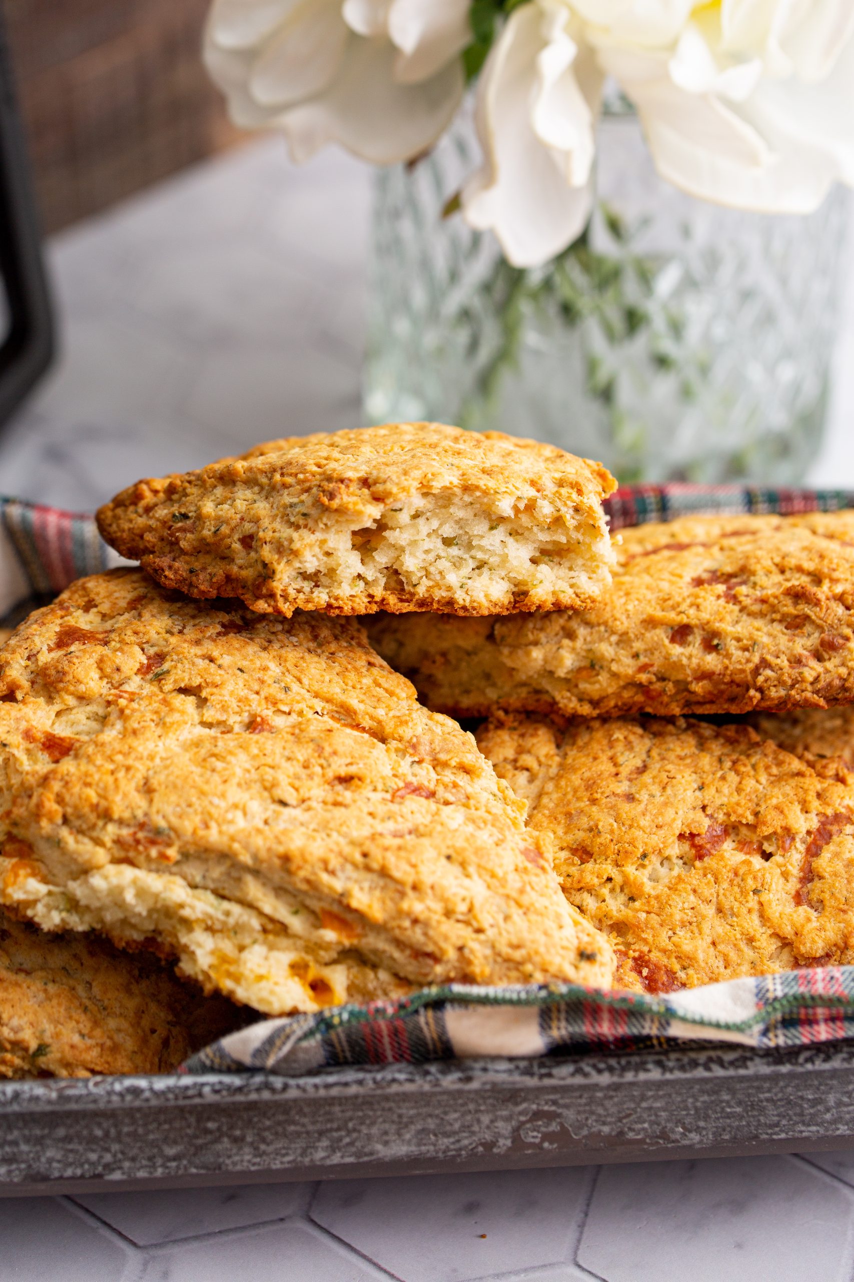 Close-up of a hand holding a half-eaten Cheddar Herb Scones, showing the crumbly interior. A tray filled with more scones and a vase of white flowers are in the background.