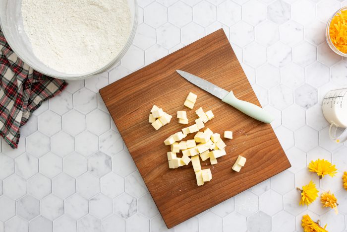 Wooden cutting board with cheese cubes and a knife. Nearby are a bowl of flour, a measuring cup with liquid, a checkered cloth, and yellow flowers on a hexagonal-tile countertop.
