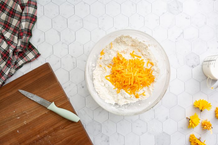 A bowl containing flour and shredded cheddar cheese on a white hexagon tile countertop, next to a knife on a wooden cutting board, flowers, and a plaid cloth.
