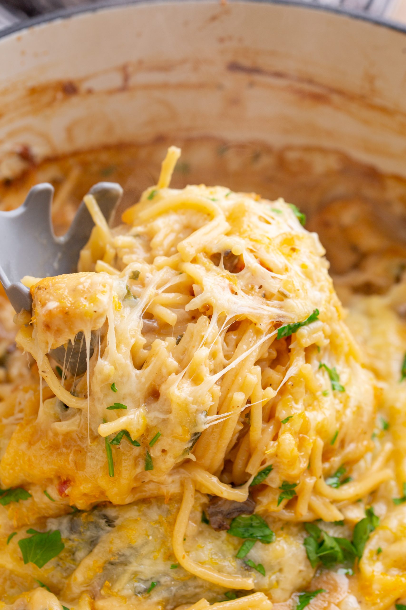 Close-up of a serving of baked cheesy pasta being lifted from a pot with a pasta fork, showing melted cheese and a garnish of fresh herbs.