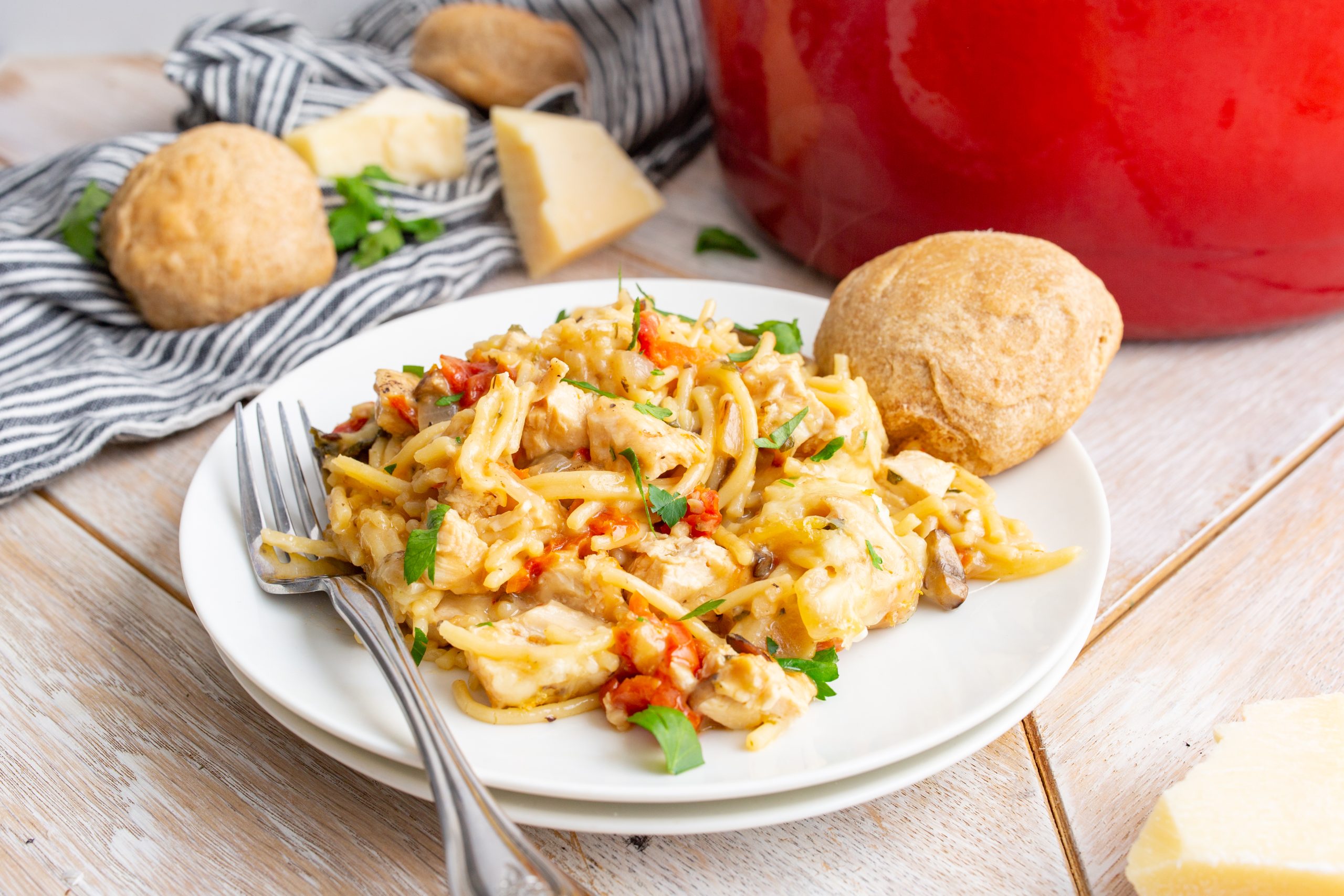 A plate of pasta mixed with vegetables and sauce, garnished with herbs. A fork rests on the plate, and a roll of bread is beside the dish. In the background, there are potatoes and a red pot.