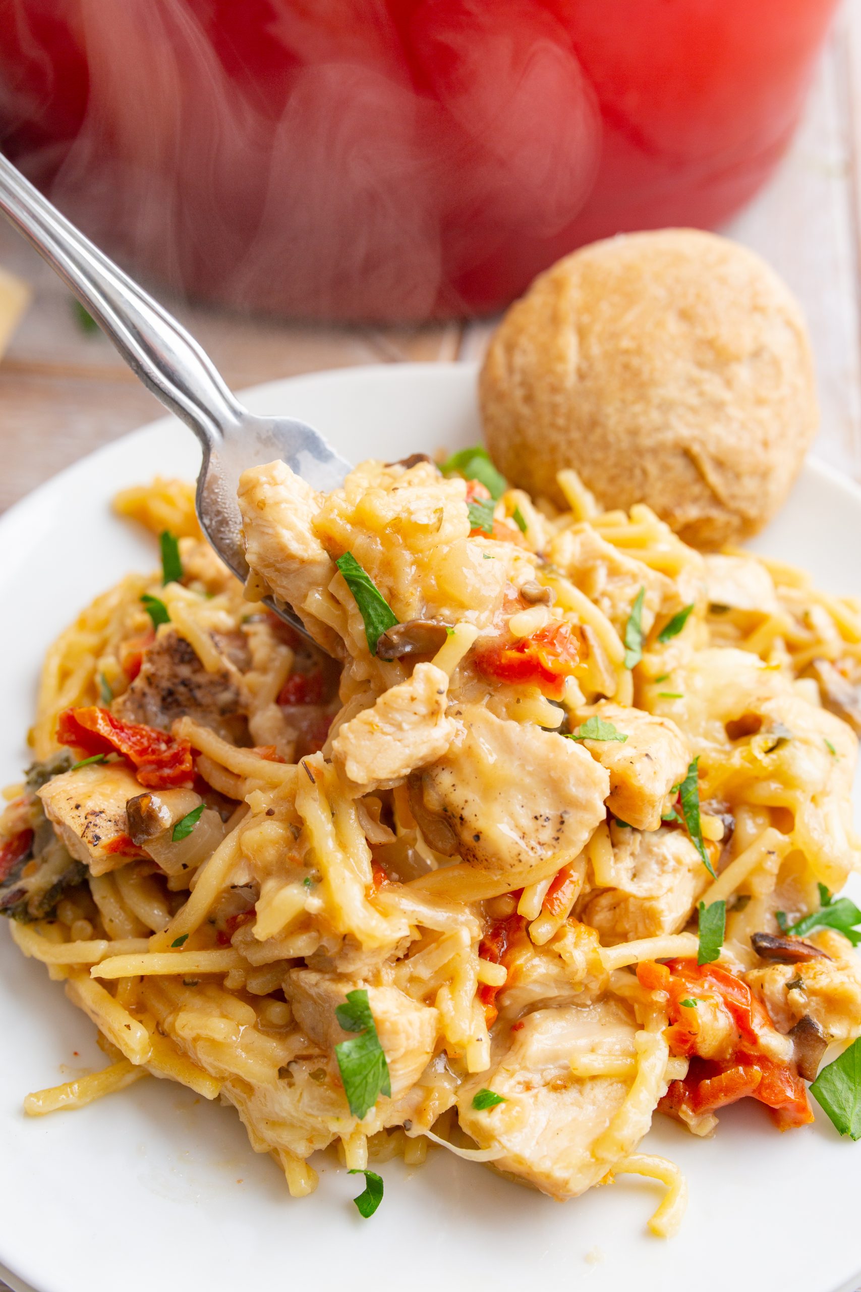 A plate of creamy chicken pasta garnished with herbs, served with a dinner roll. A fork is lifting a bite of pasta and chicken.