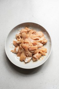 Raw, marinated chicken pieces in a white bowl on a light grey surface.