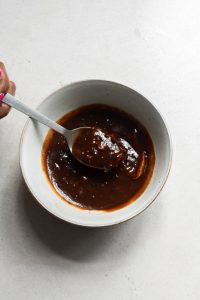 A person's hand holding a spoon, stirring a bowl of dark brown, thick sauce on a light gray surface.