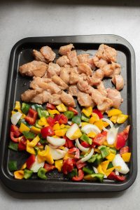 Raw diced chicken and chopped colorful bell peppers with onions on a baking tray, ready for cooking.