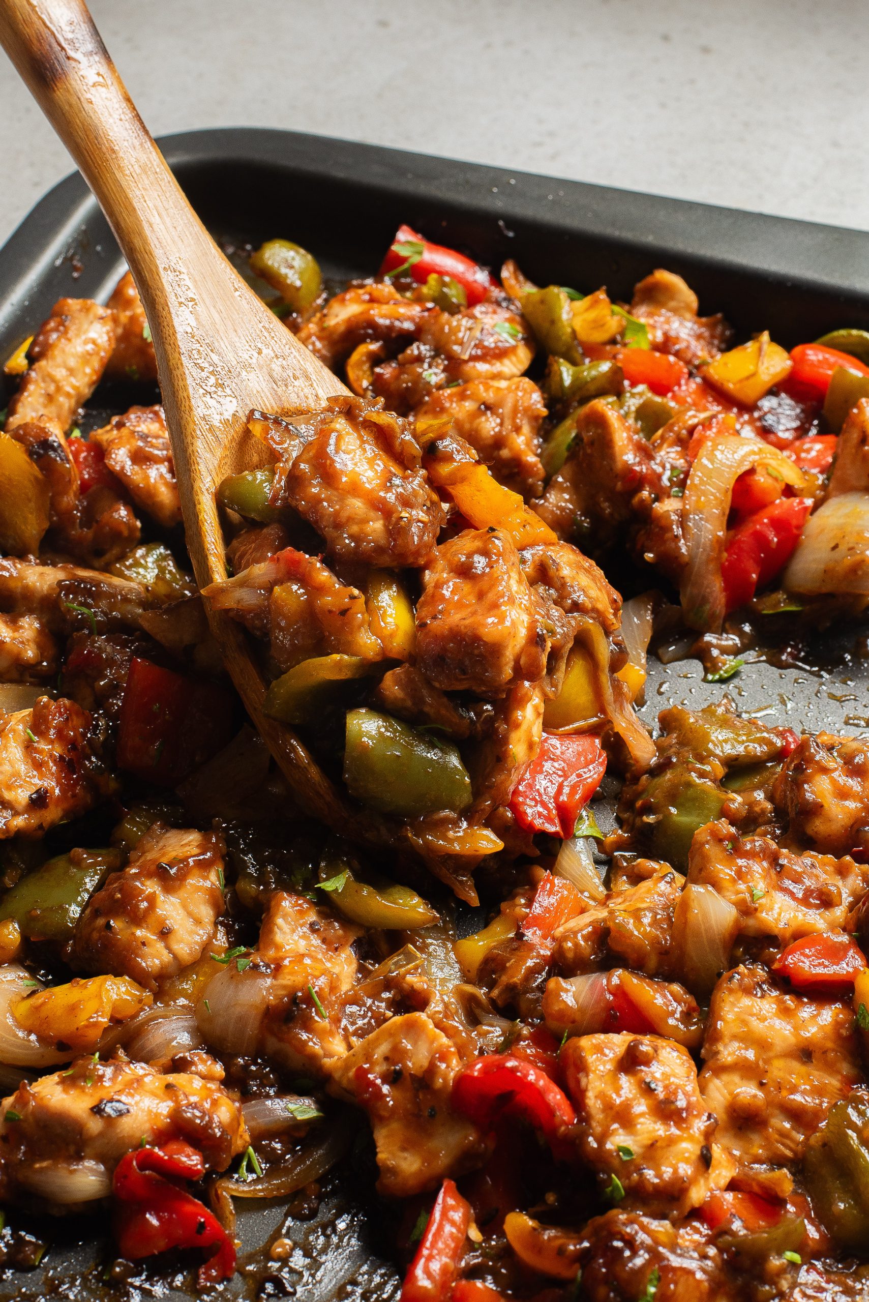 A skillet filled with stir-fried chicken and vegetables including bell peppers and onions, with a wooden spoon.