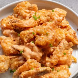A white plate filled with crispy fried shrimp covered in a creamy, orange sauce, garnished with chopped green onions.