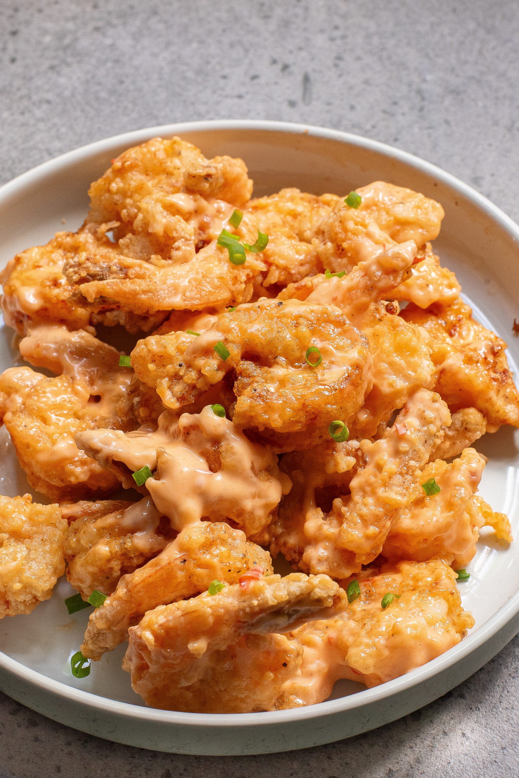 A white plate filled with bang bang shrimp covered in a creamy, orange sauce, garnished with chopped green onions.