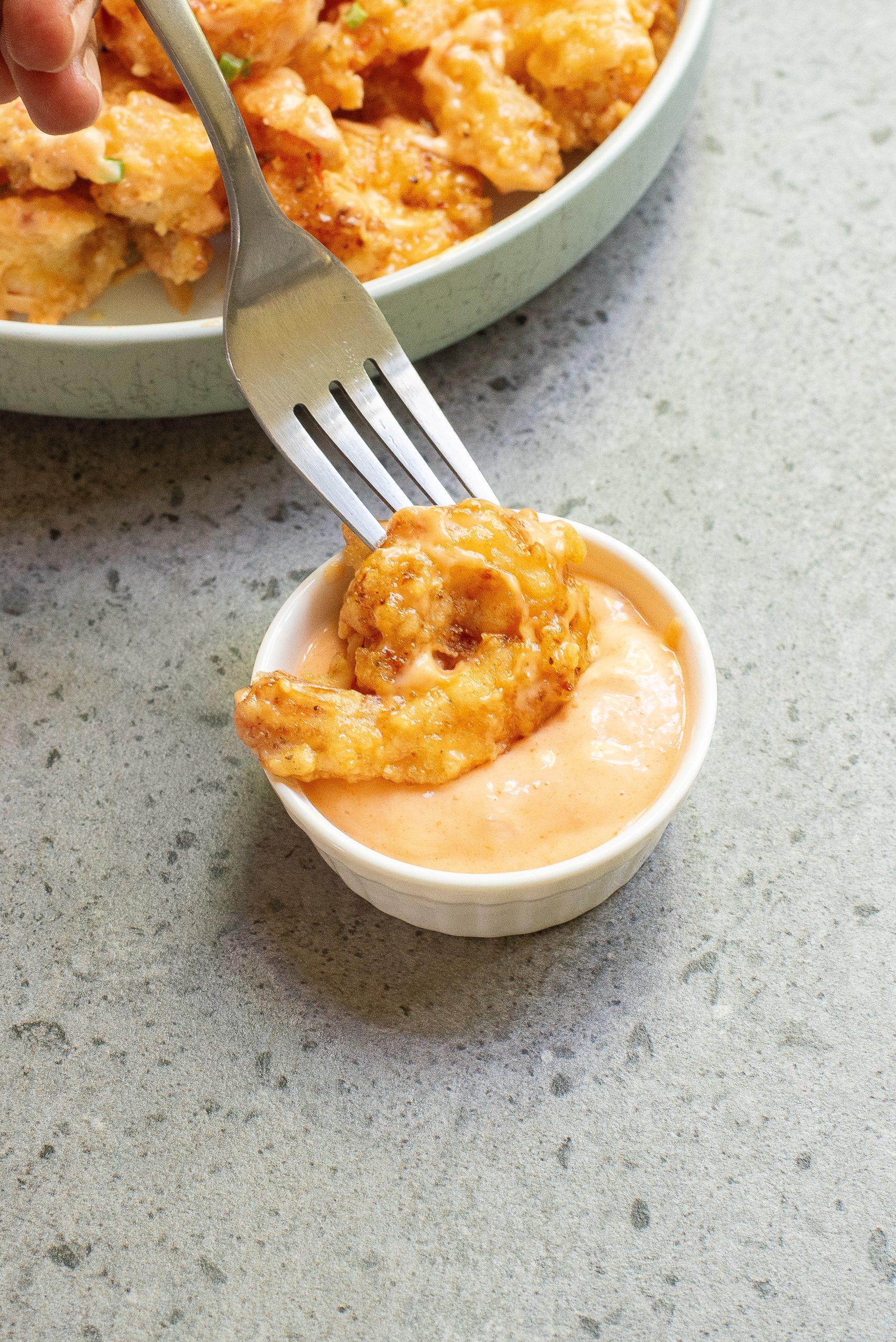 A fork dips a piece of breaded shrimp into a small bowl of creamy orange sauce on a gray surface, with a plate of more shrimp in the background.