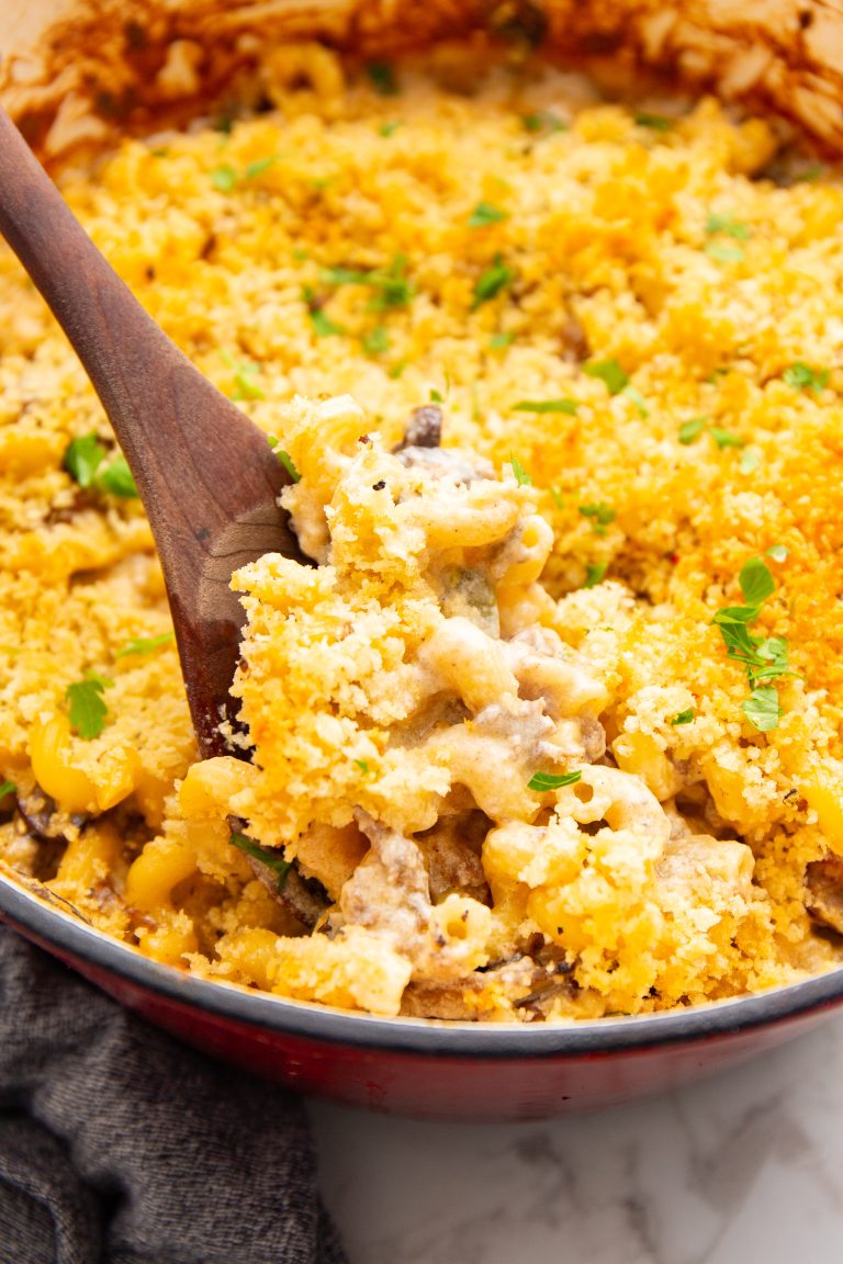 Close-up of a wooden spoon scooping cheesy pasta bake topped with breadcrumbs from a red casserole dish. The dish contains creamy pasta mixed with vegetables, garnished with chopped herbs.