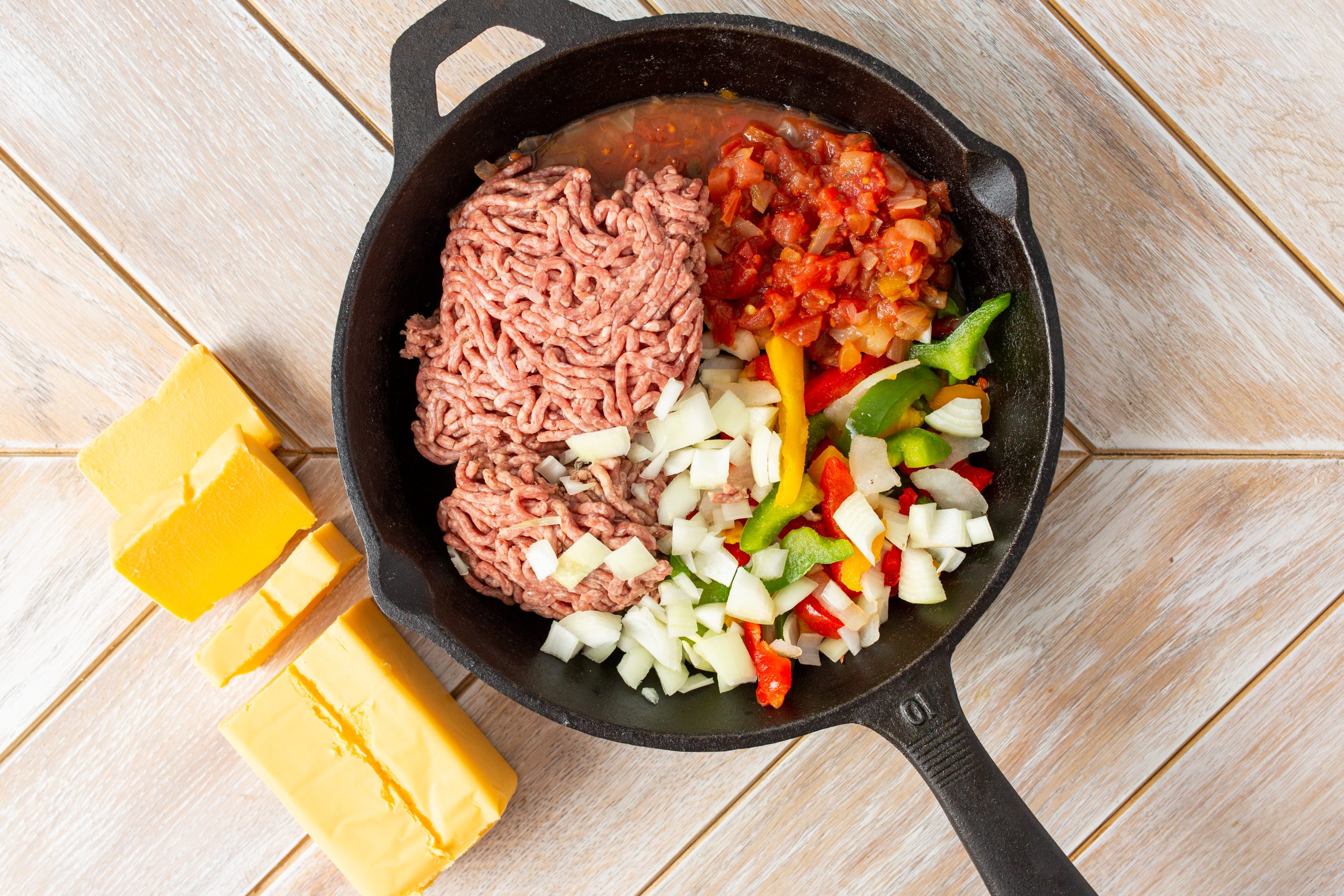 A cast iron skillet holds ground beef, diced onions, sliced bell peppers, and chopped tomatoes. Next to the skillet are three blocks of yellow cheese on a wooden surface.