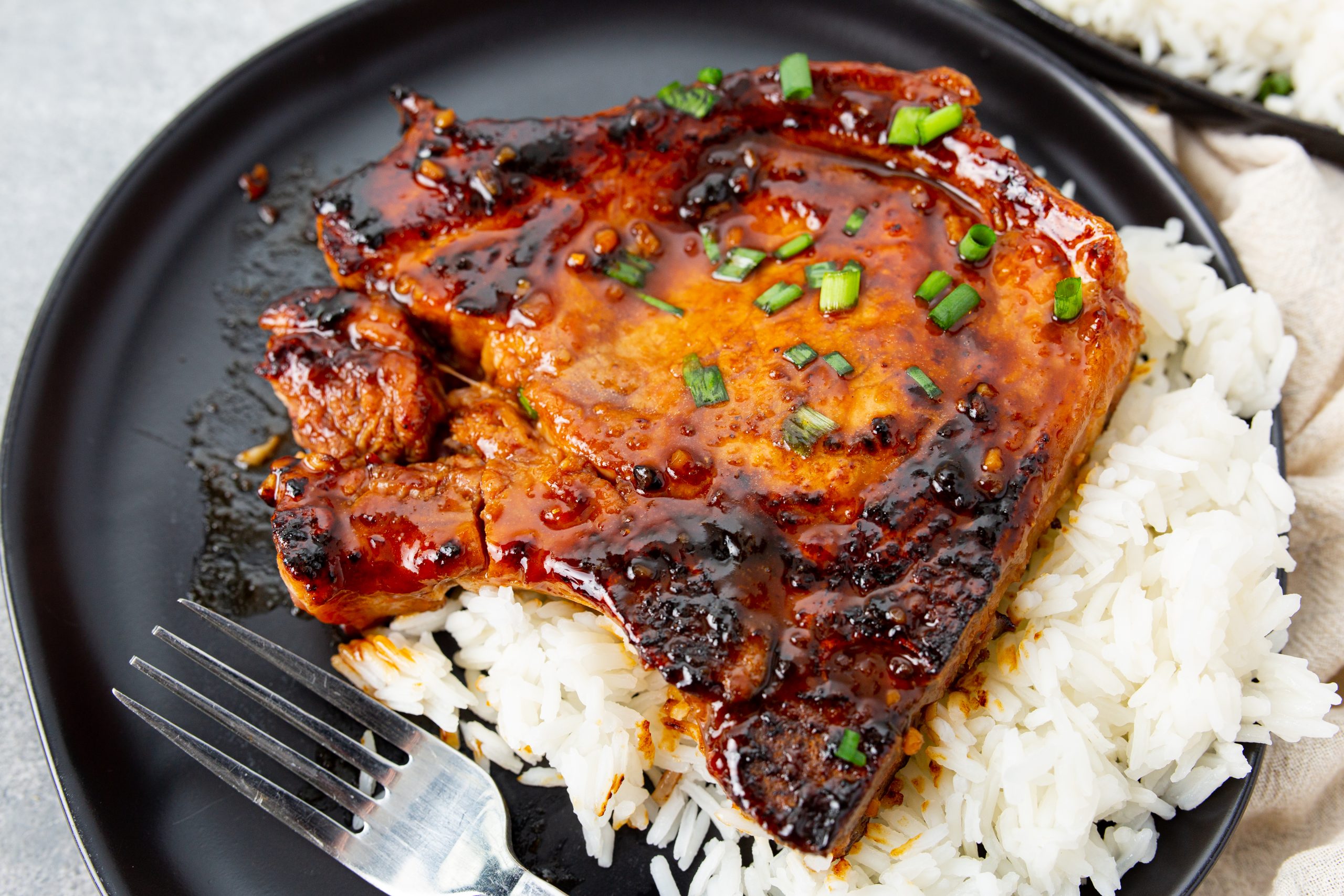 A grilled pork chop glazed with sauce is served on white rice with chopped green onions on a black plate, next to a fork.