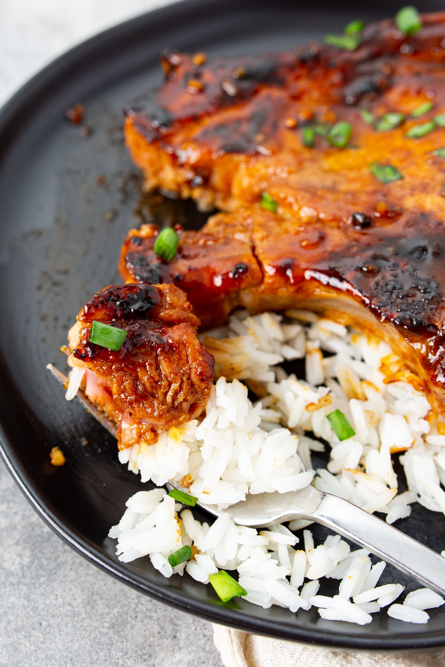 A grilled pork chop with a bite taken out is placed on a plate over white rice, garnished with green onions. A fork rests on the side of the plate.