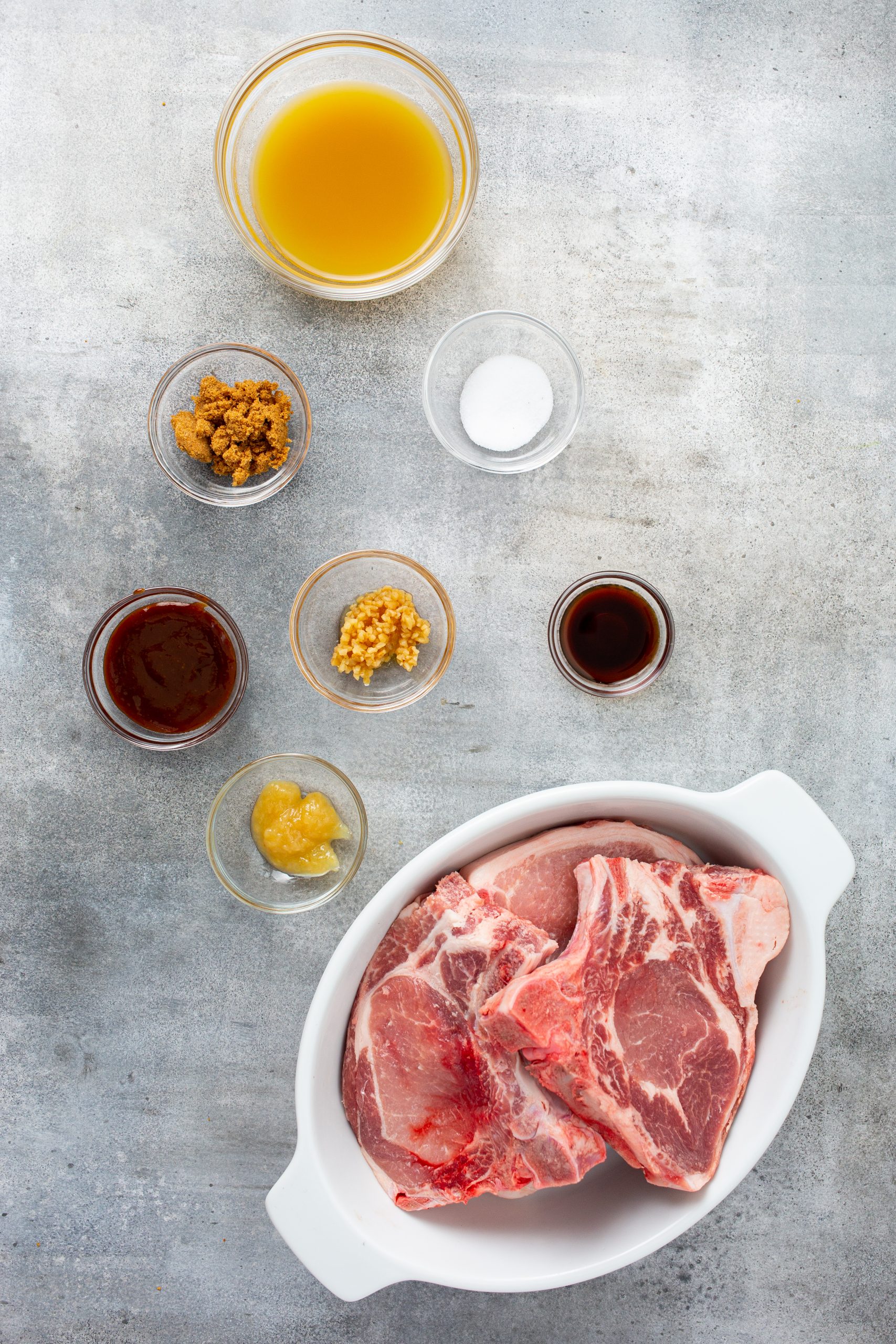 A white dish with raw pork chops, surrounded by bowls containing various ingredients: broth, brown sugar, salt, minced garlic, sauce, and mustard, arranged on a gray countertop.
