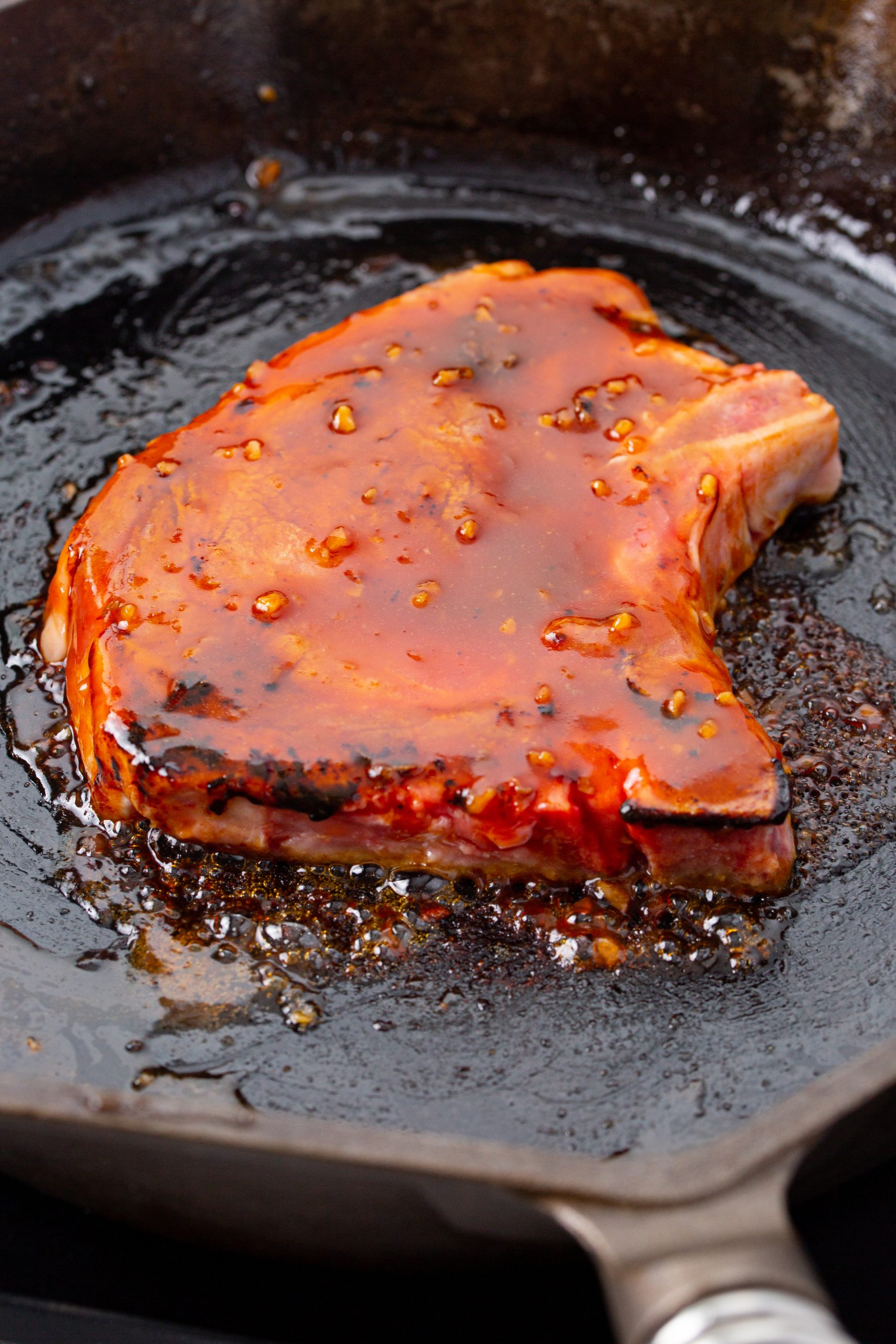 Meat with a reddish glaze is cooking in a cast iron skillet. The glaze is bubbling and caramelizing around the edges of the meat.