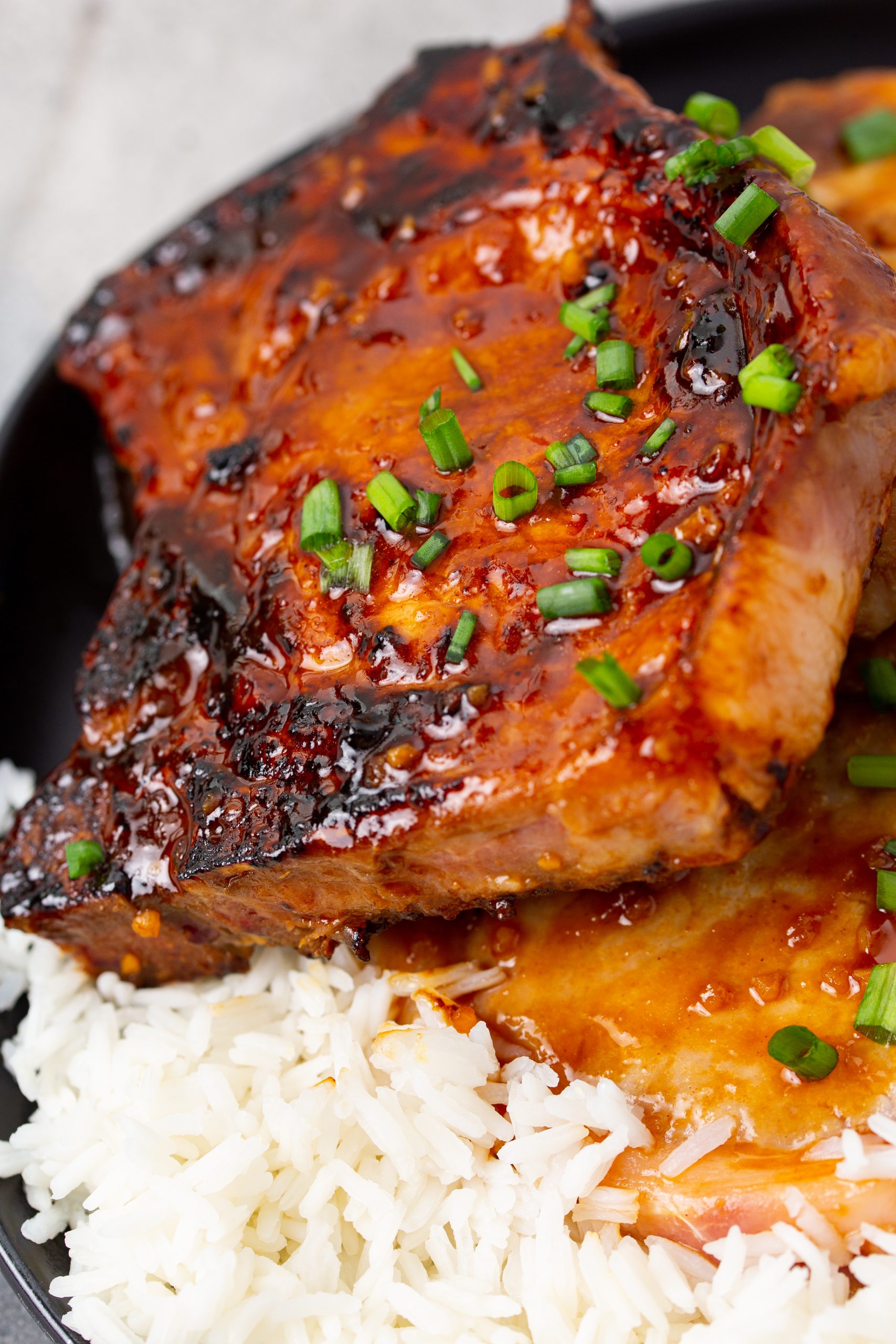 A grilled pork chop glazed with sauce is served on white rice with chopped green onions on a black plate, next to a fork.