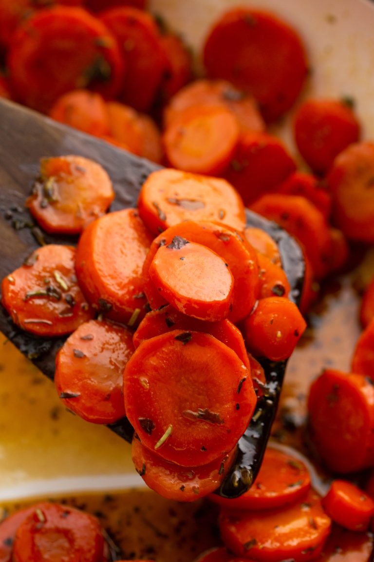 Close-up of a wooden spatula with glazed, sliced carrots garnished with herbs. More sliced carrots are visible in the background.