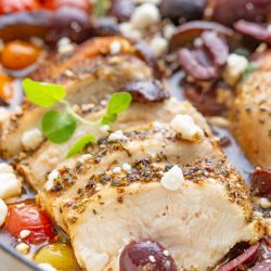 Sliced, seasoned chicken breast is garnished with olives, tomatoes, and crumbled feta cheese. A sprig of greenery adds a final touch to the dish.