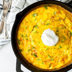 A baked dish with a golden crust in a black skillet, topped with a dollop of sour cream, and garnished with green herbs. A white and black patterned cloth and three forks are beside the skillet.