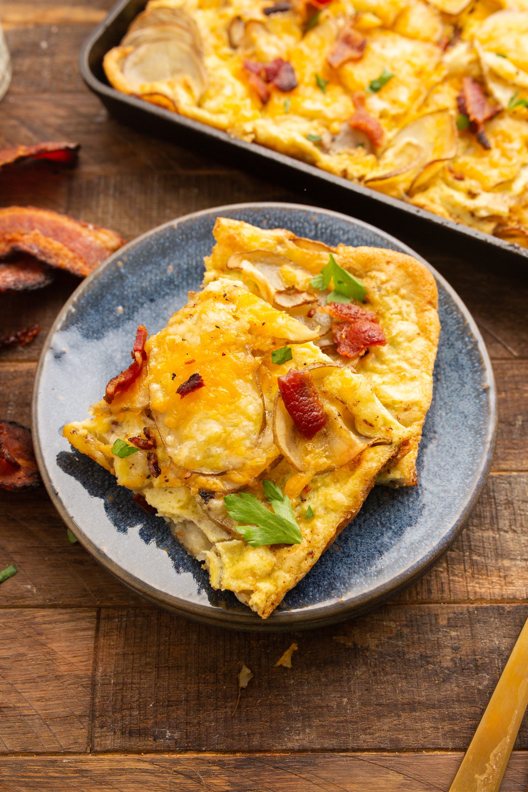 A slice of potato and bacon breakfast pizza on a blue plate next to a baking sheet with more pizza. The pizza has a golden crust, melted cheese, and garnished with parsley. A knife and bacon strips are nearby.