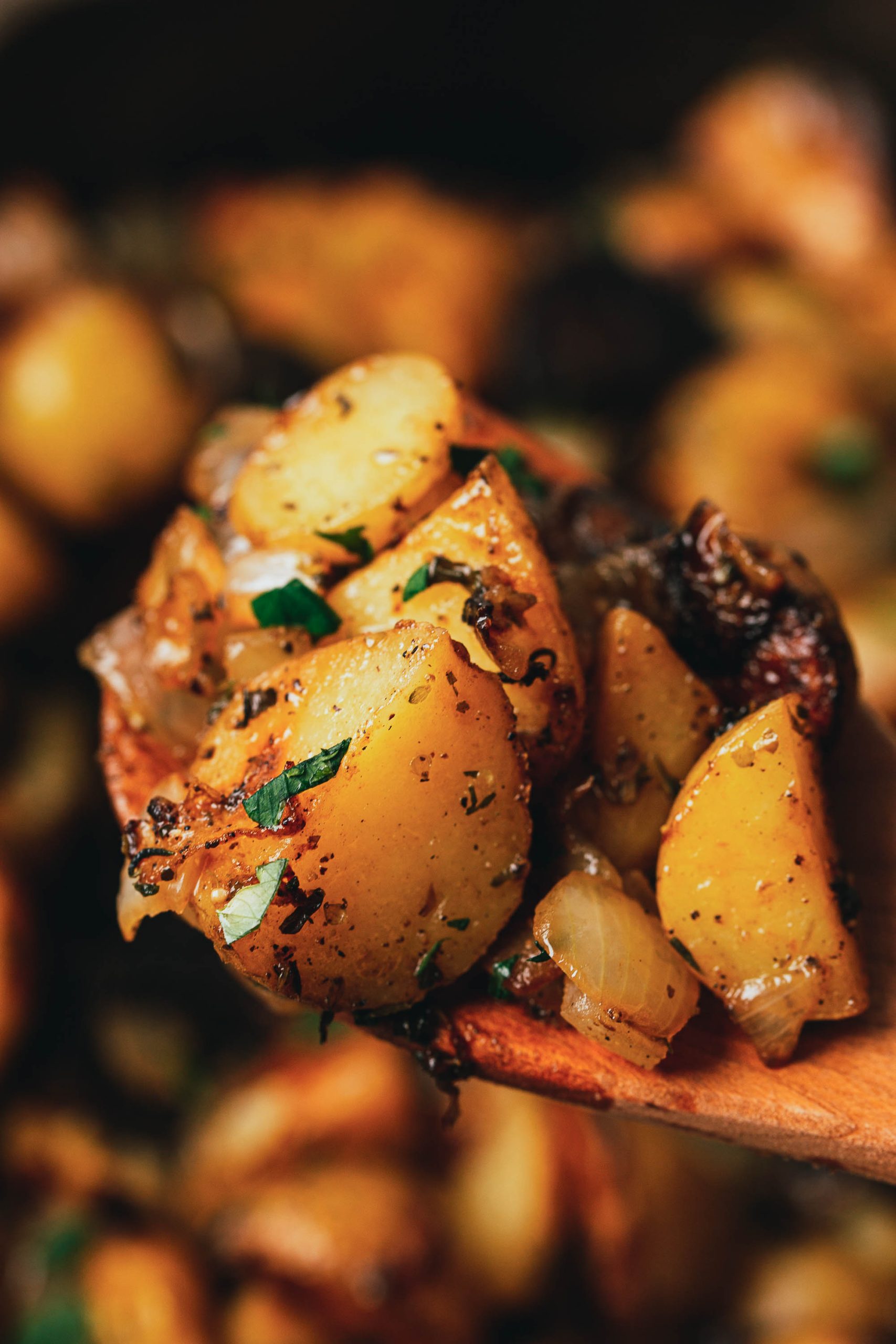 Close-up of roasted potatoes with charred edges, garnished with chopped herbs, being scooped with a wooden spoon.