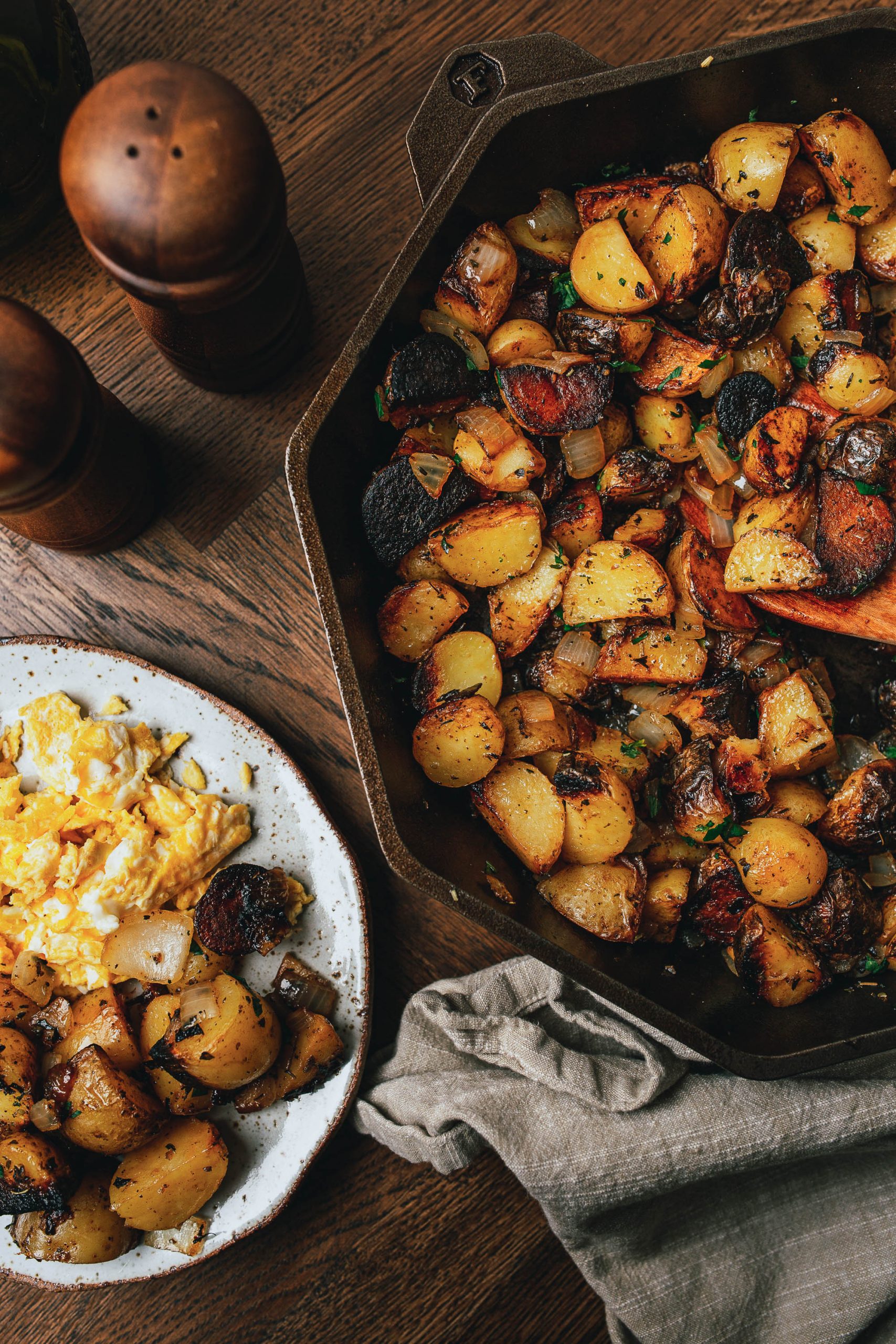 A skillet filled with cooked potatoes and vegetables is on a wooden table. A separate plate with scrambled eggs and a serving of the potato dish is next to it. Salt and pepper shakers are nearby.