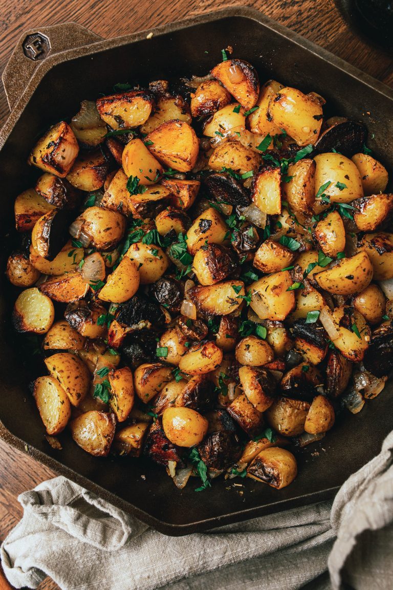 A cast iron pan filled with roasted diced potatoes, mushrooms, and onions, garnished with chopped parsley. A cloth napkin is partially visible underneath the pan.