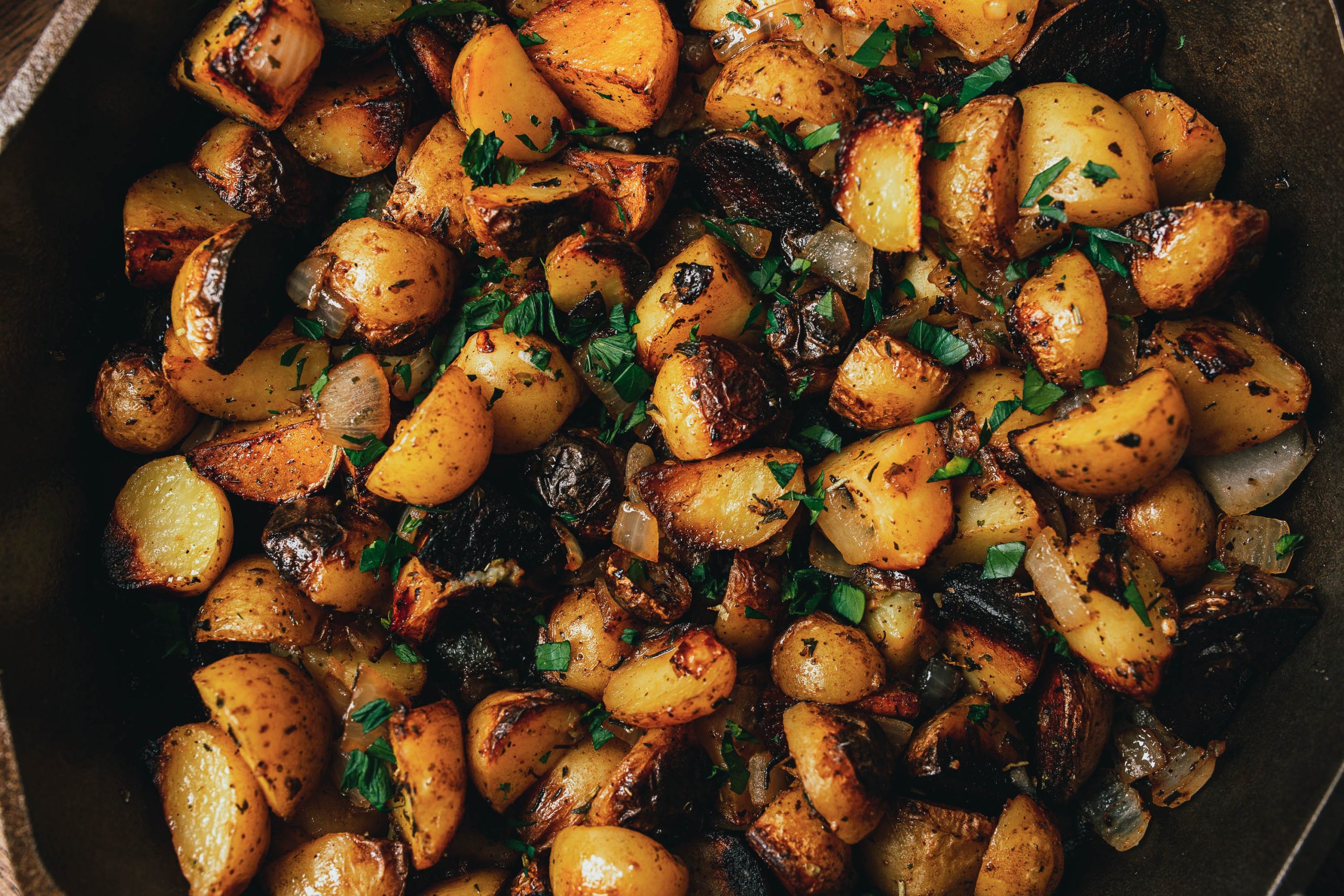 A close-up of seasoned roasted potatoes with charred edges, garnished with chopped fresh herbs. The potatoes appear crispy and golden brown.