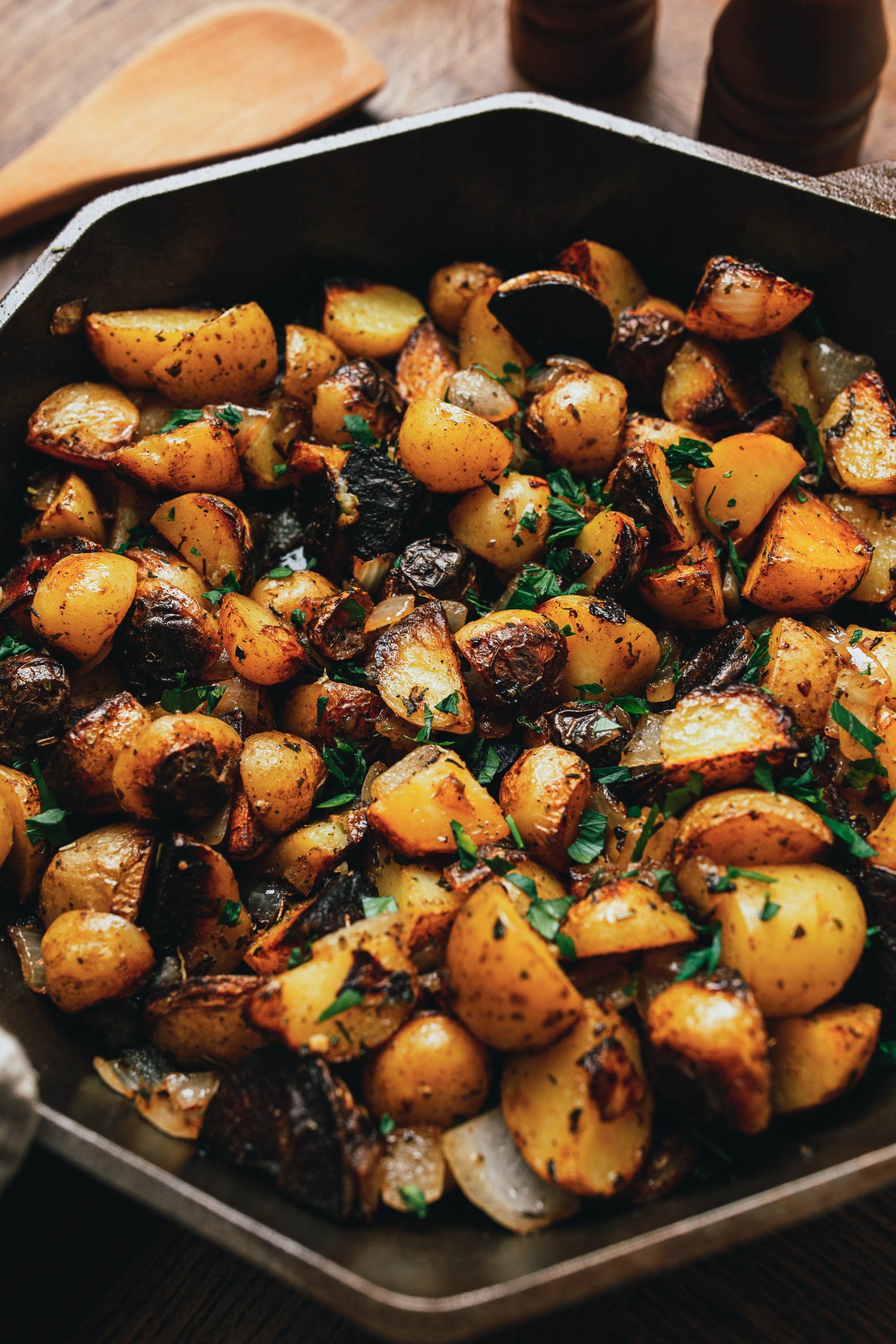 A skillet filled with roasted baby potatoes, chunks of garlic, and herbs, placed on a wooden table with salt and pepper mills in the background.