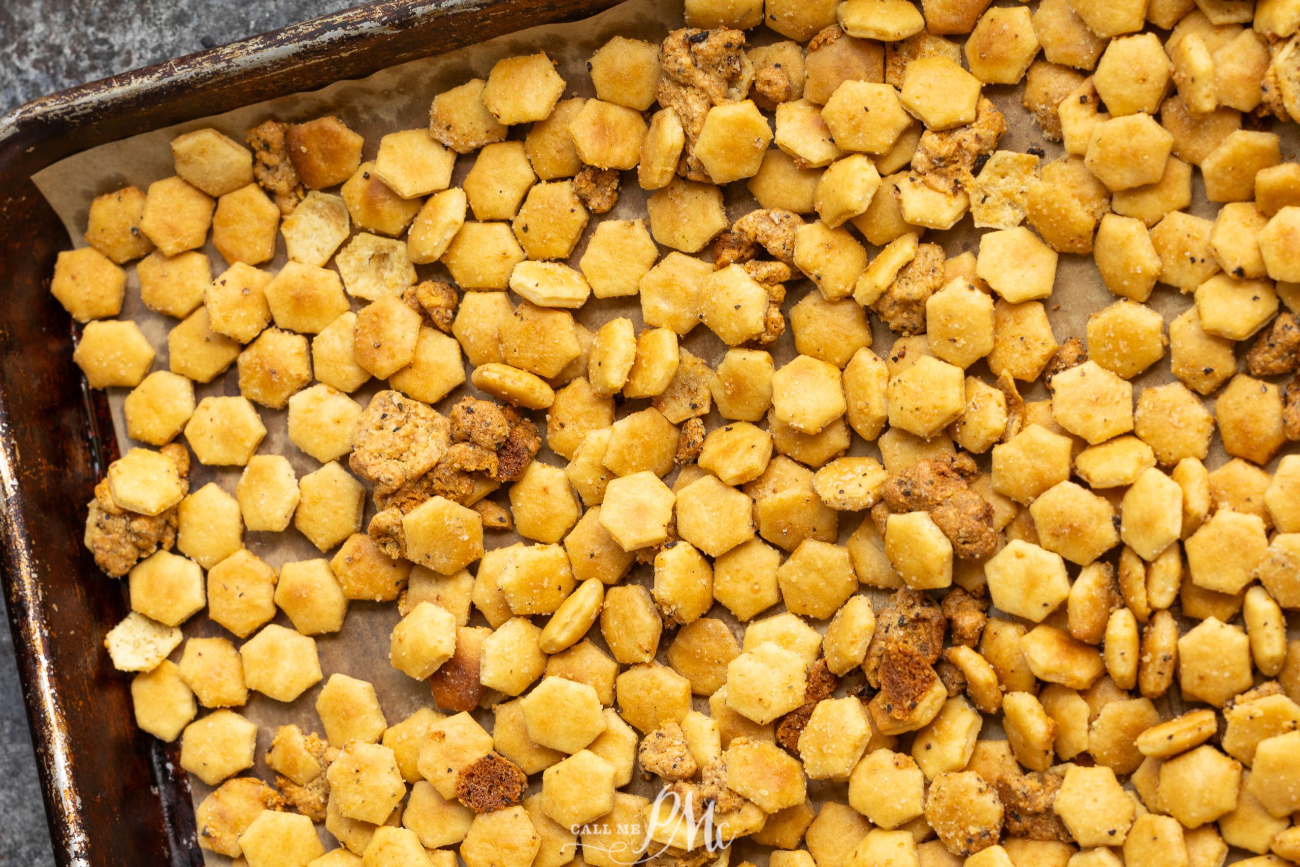 A baking sheet is filled with small, hexagonal crackers and bits of other small snacks, all toasted to a golden-brown color. The edges of the baking sheet are slightly charred.