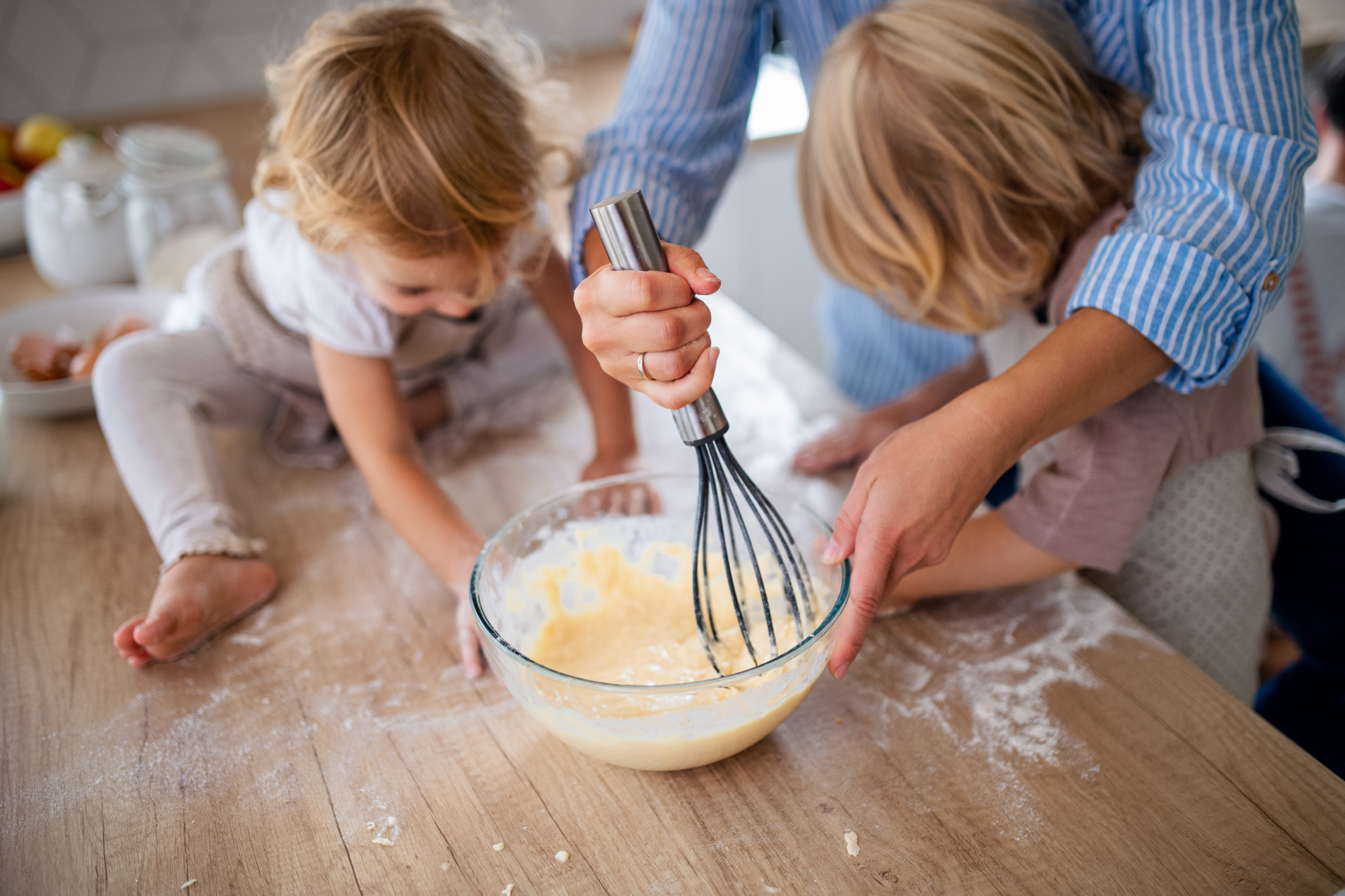 Two young kids mix batter in a glass bowl on a flour-dusted kitchen counter, focusing intently on their cooking task.
