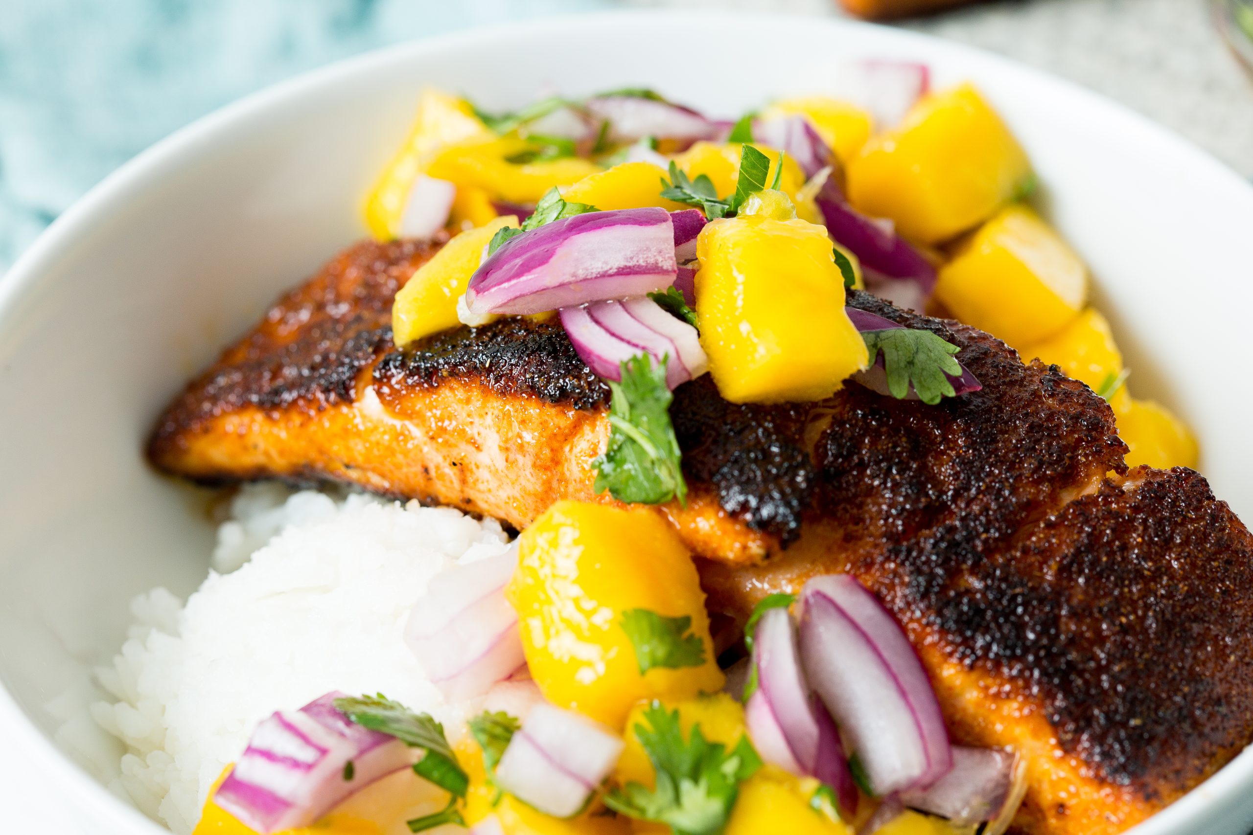 A bowl with a piece of blackened salmon topped with a colorful mango salsa and served over white rice.