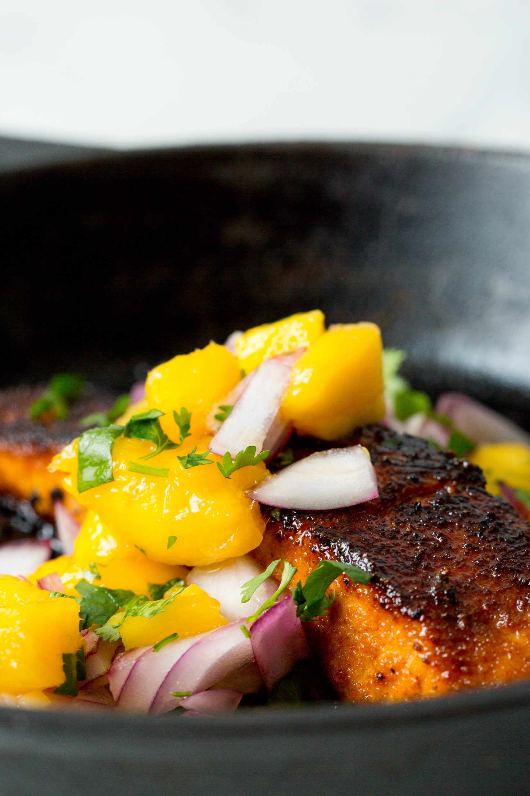 A close-up of a blackened piece of fish topped with a fresh mango salsa, featuring diced mango, red onion, and cilantro, served in a black dish.